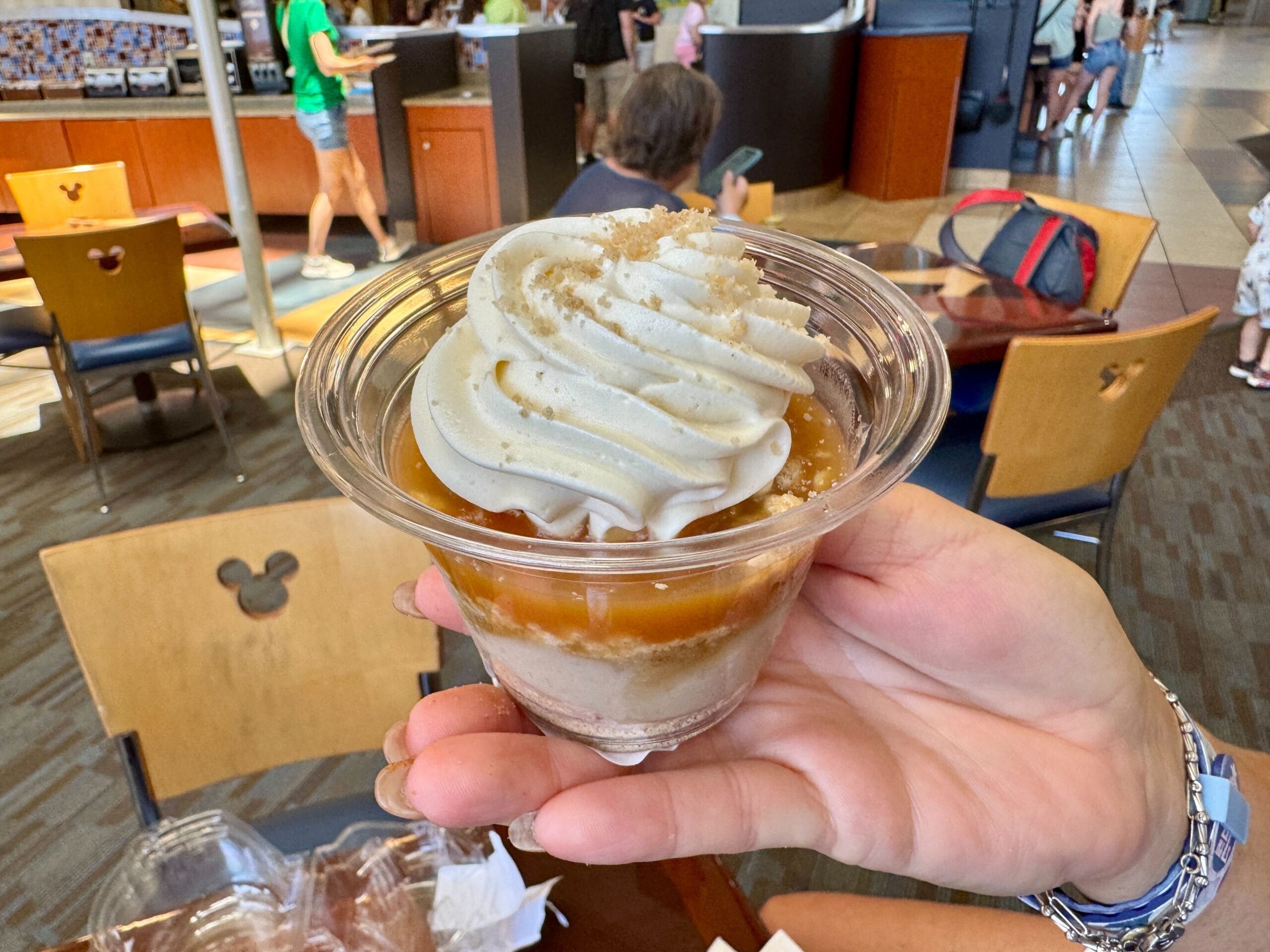 A hand holds a dessert cup with whipped topping and caramel sauce at Contempo Cafe, surrounded by wooden chairs.