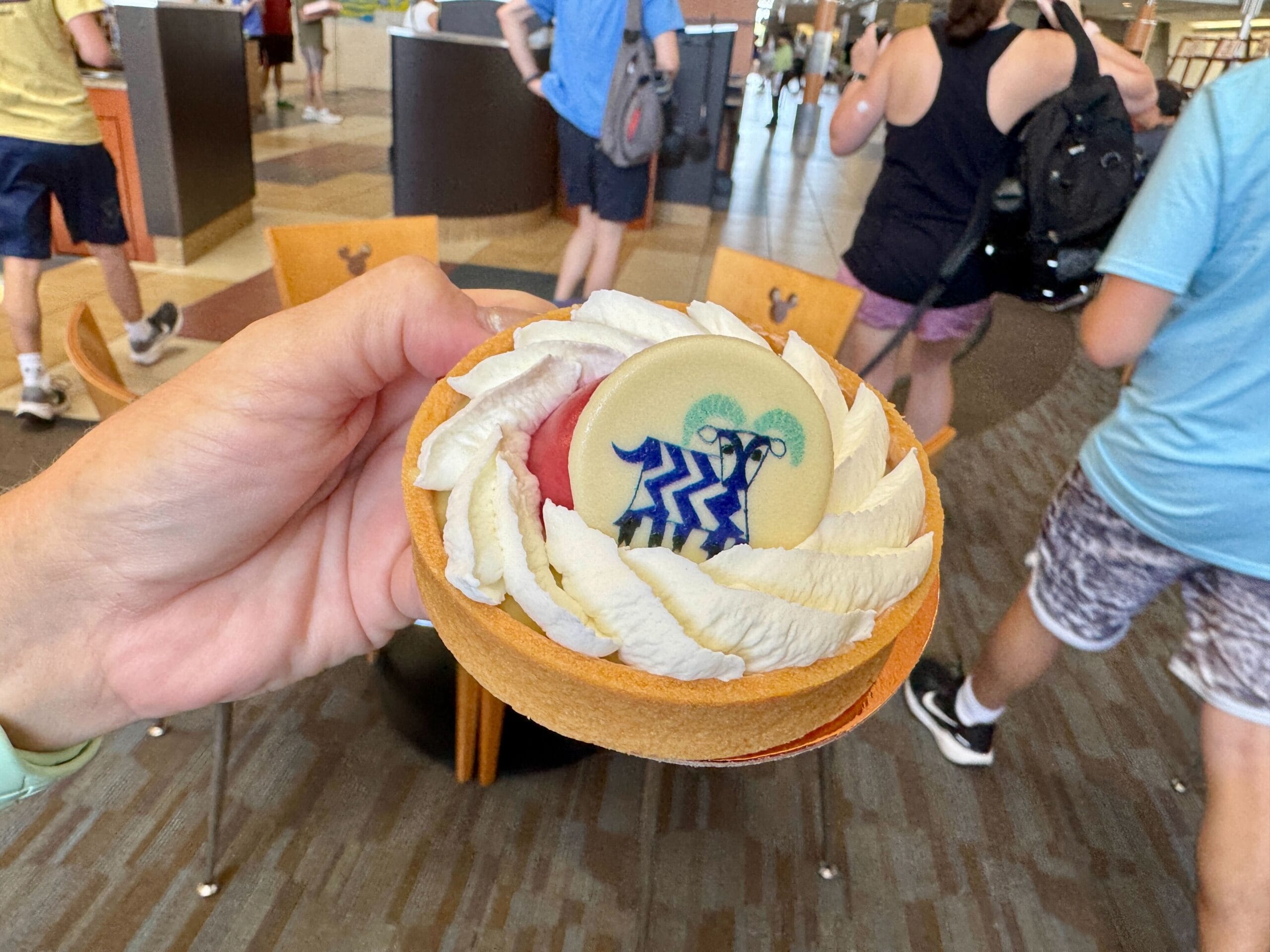 A hand holds a Contempo Cafe tart topped with cream and a blue animal chocolate disc, indoors with people walking.