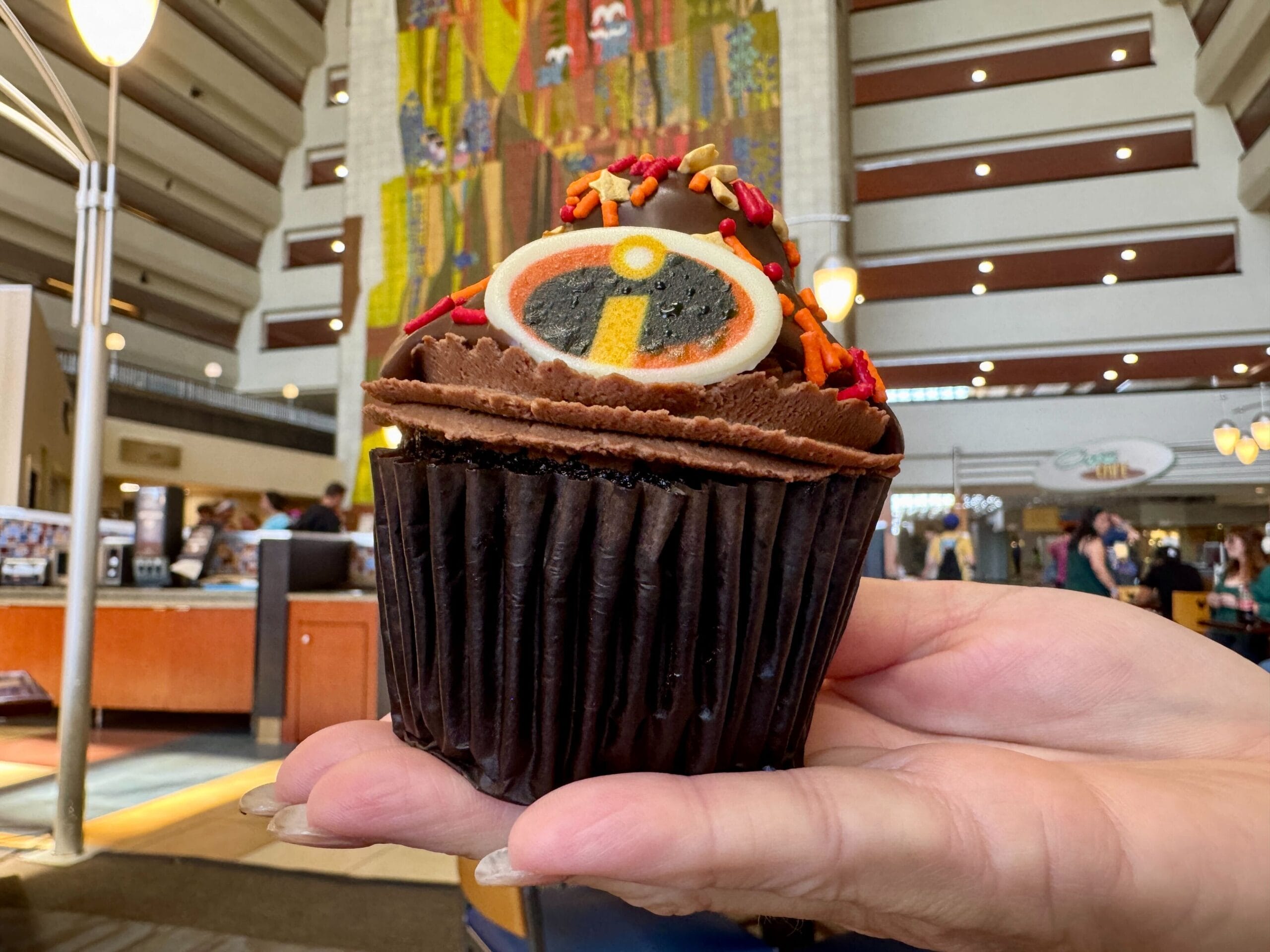 A hand holds a chocolate cupcake with The Incredibles logo at Contempo Cafe, known for desserts and breakfast pastries.