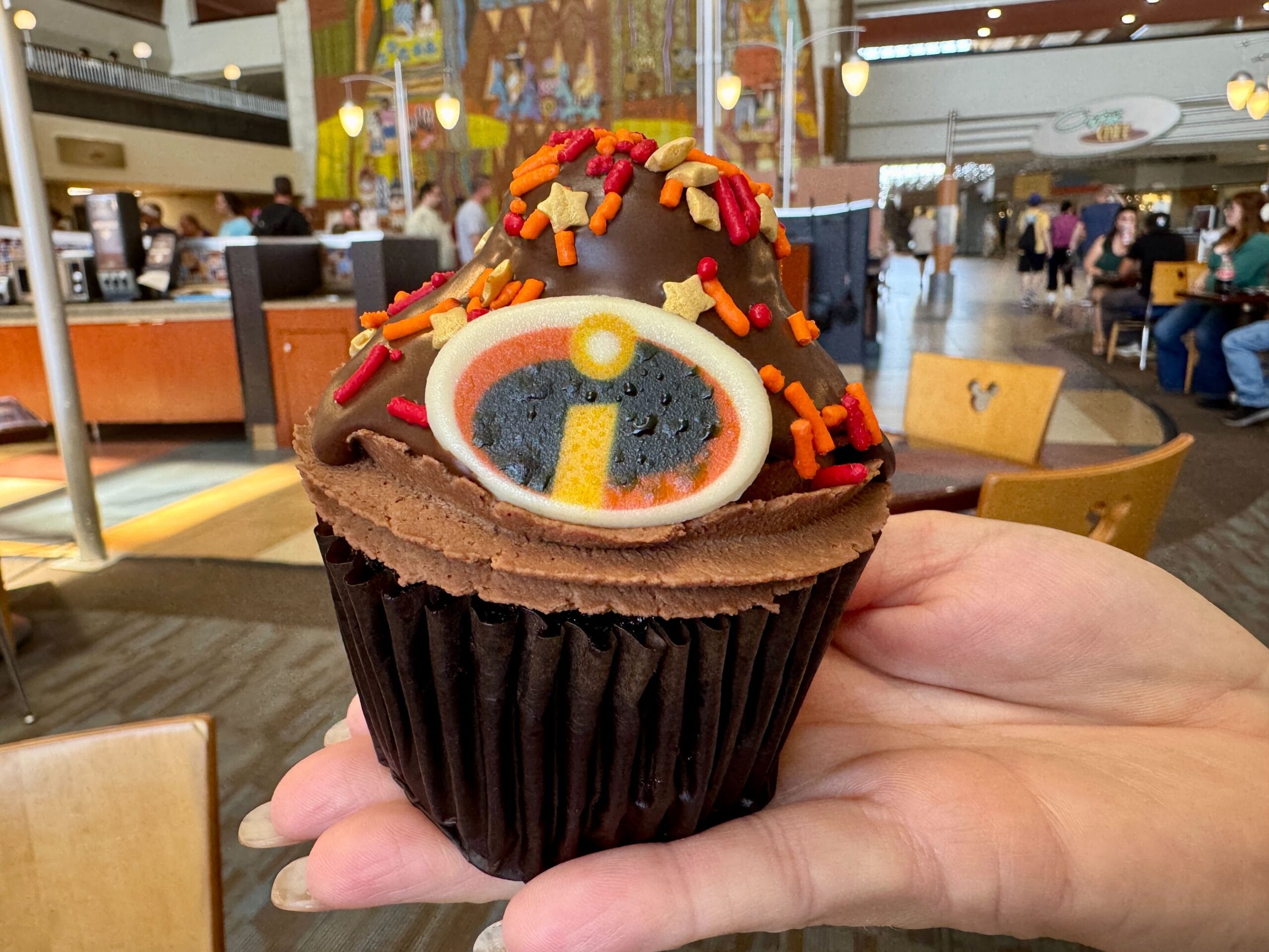 A hand holds a chocolate cupcake from Contempo Cafe topped with The Incredibles logo and vibrant sprinkles—Desserts delight!