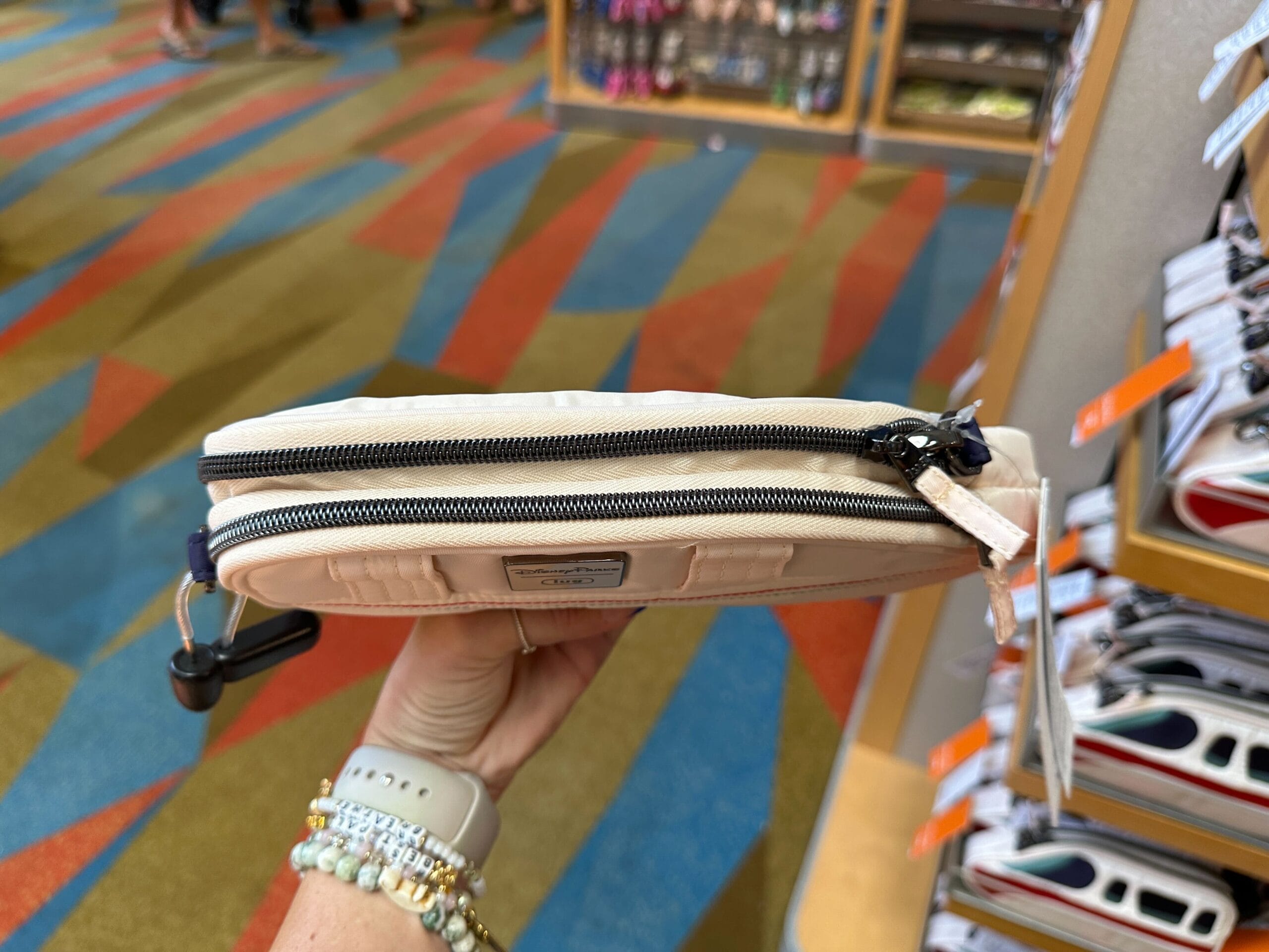A hand holds a zippered beige pouch in a store with colorful carpet and shelves in the background.