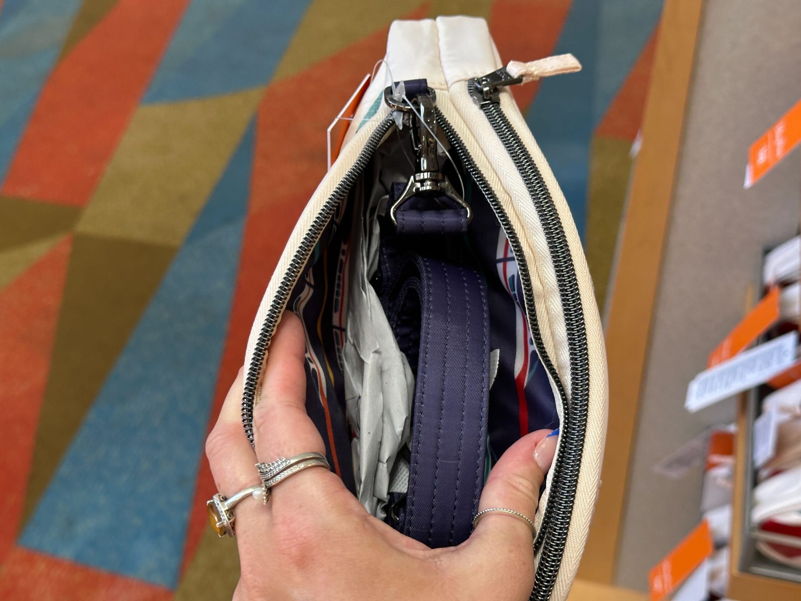 Hand holding an open zippered bag with a strap inside, over a colorful patterned carpet.