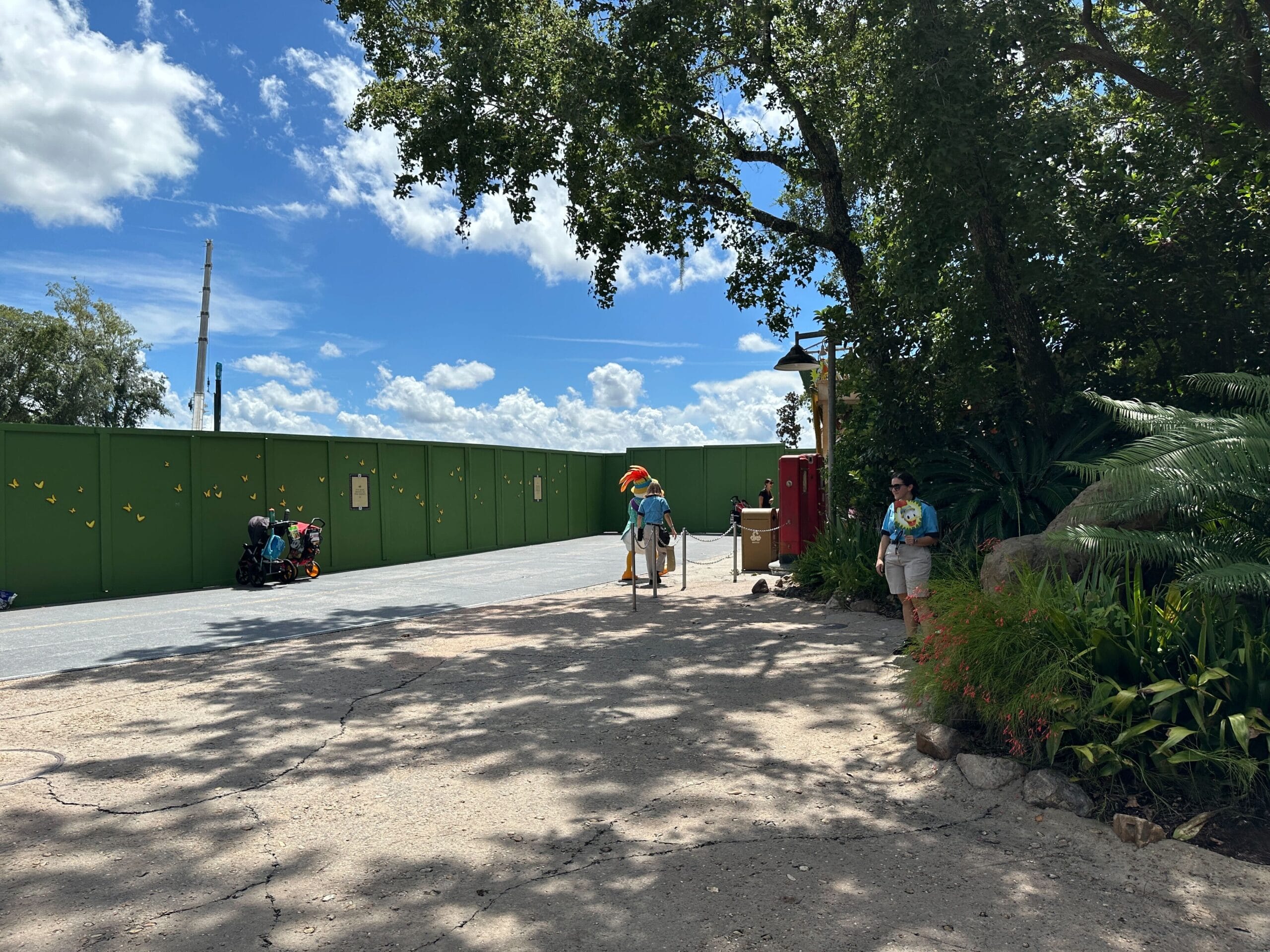 People walk by green construction walls in DinoLand U.S.A. in Walt Disney World under a sunny, partly cloudy sky.