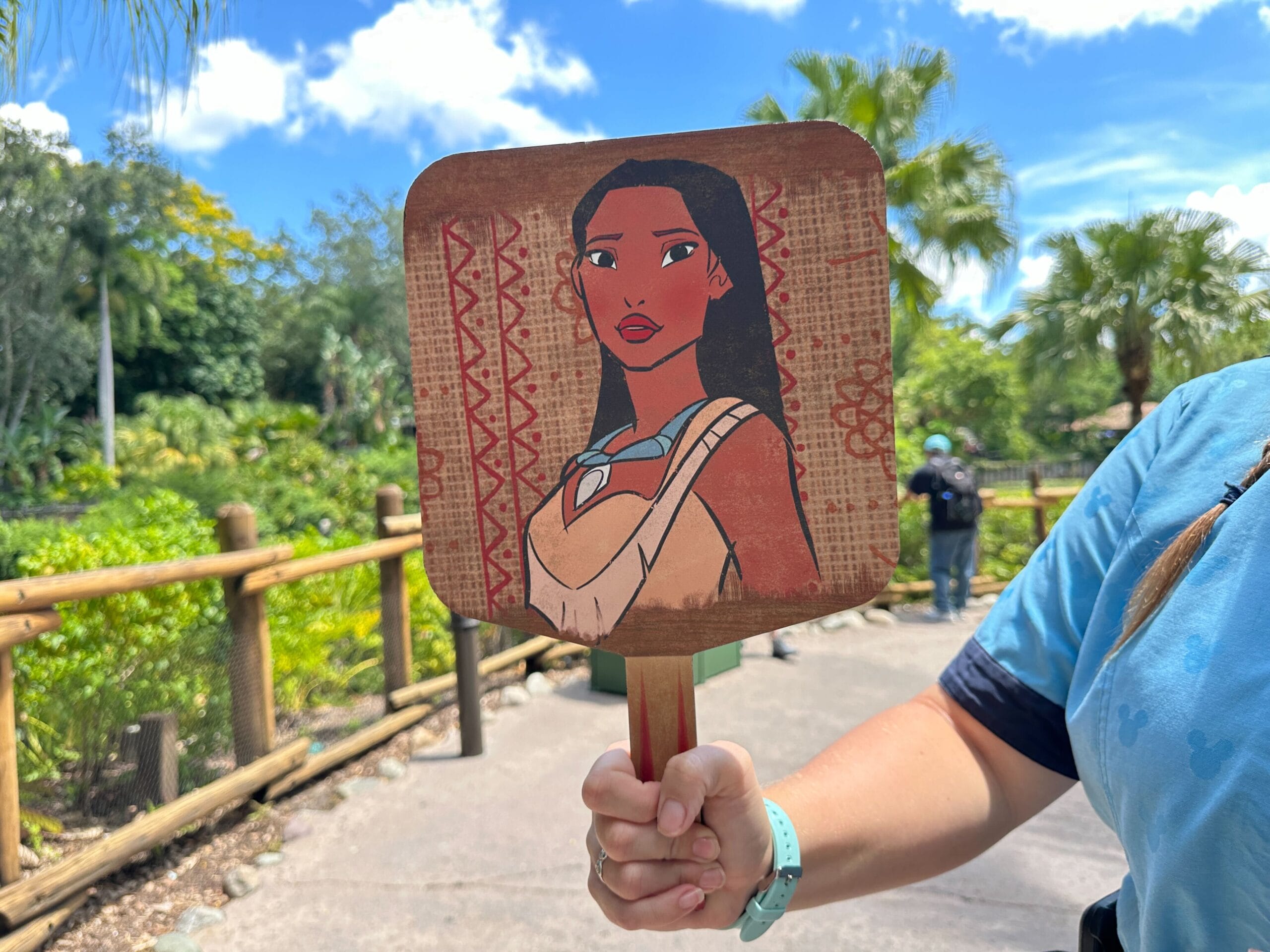 A person holds a Walt Disney World fan with Pocahontas outdoors on a sunny day, trees and blue sky in the background.