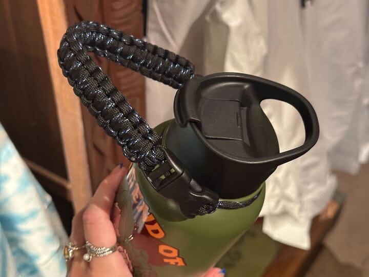 A hand holds a green water bottle with a black paracord handle attached to the lid.