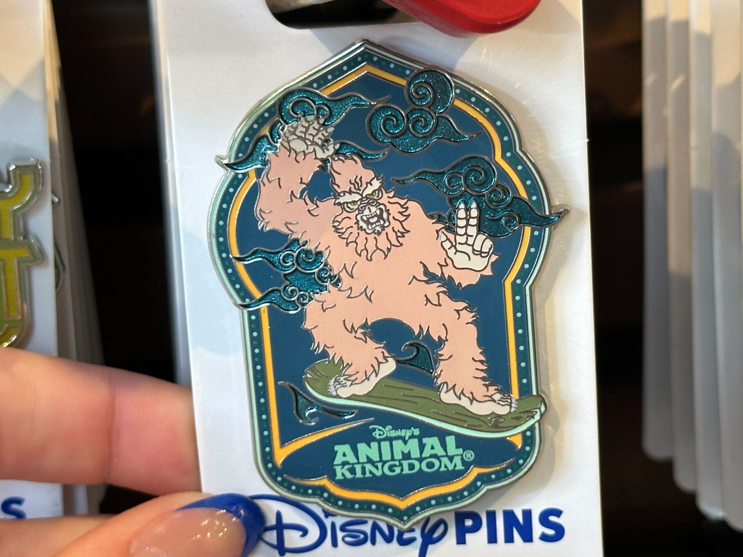A Disney's Animal Kingdom pin featuring a pink yeti surfing on a snowboard, held by a person with blue-painted nails.