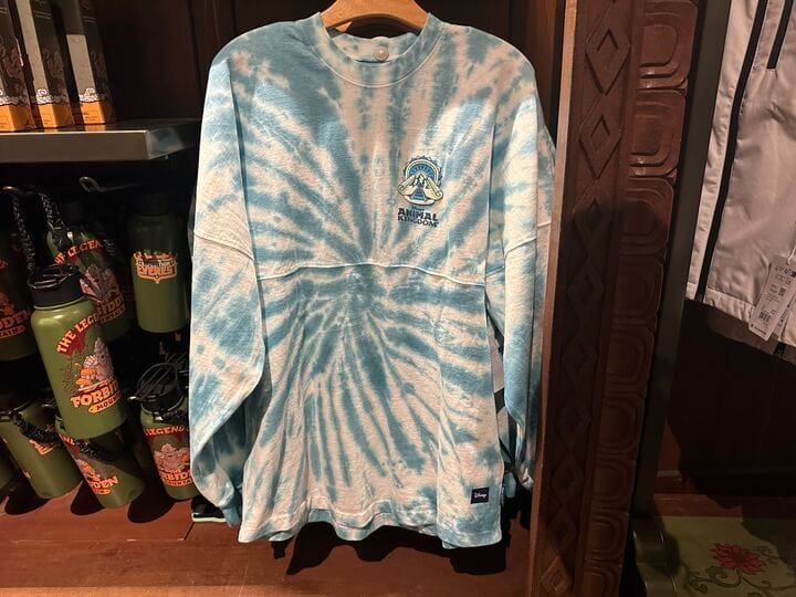 A blue and white tie-dye long-sleeve shirt with a small Animal Kingdom logo hangs on a wooden hanger in a store.