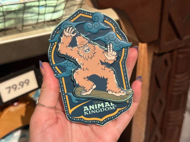 A hand holds an Animal Kingdom sticker featuring a surfing yeti with blue clouds in the background.