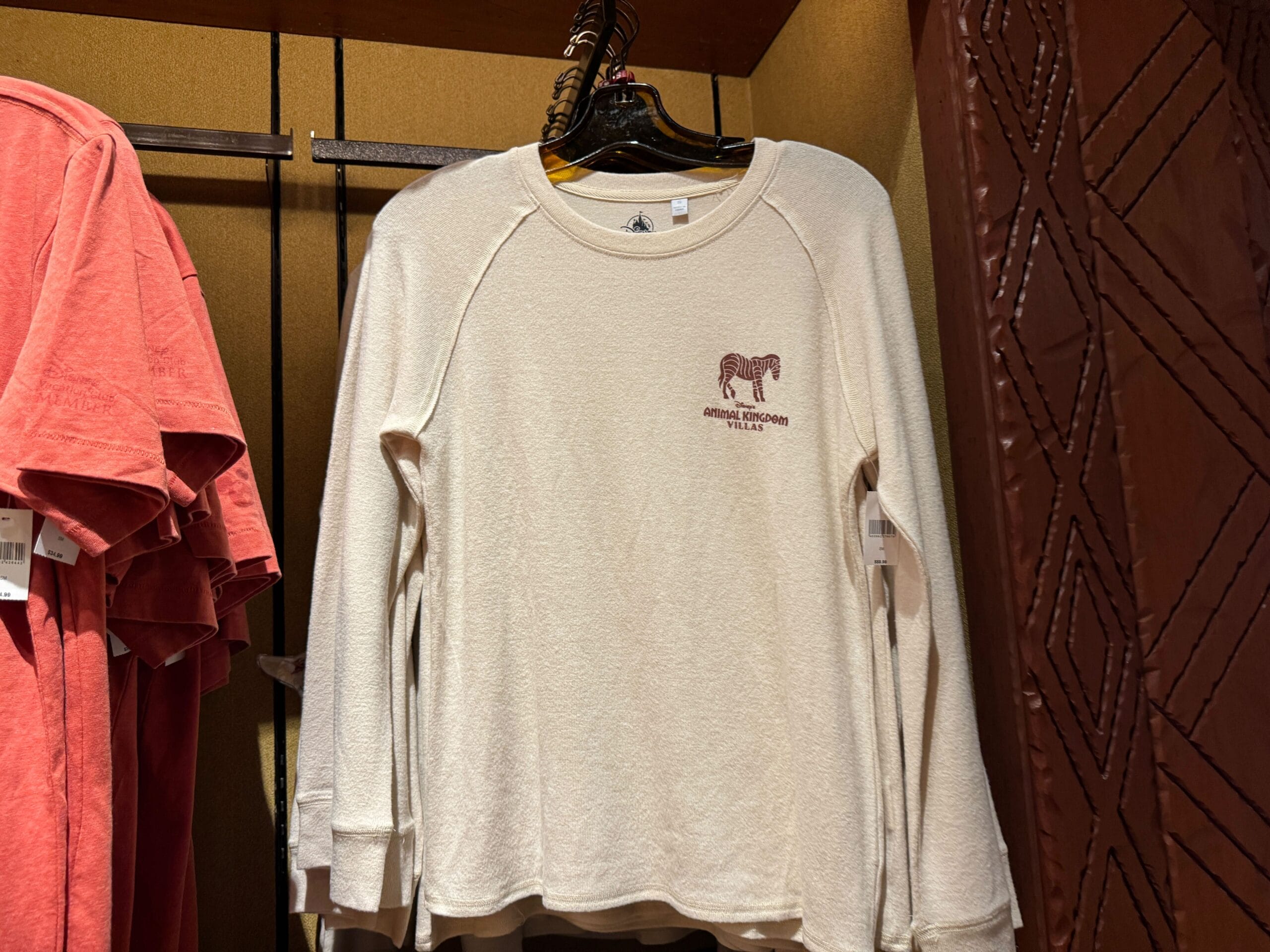 Cream long-sleeve shirt with a small zebra graphic and text hanging on a rack beside coral shirts.