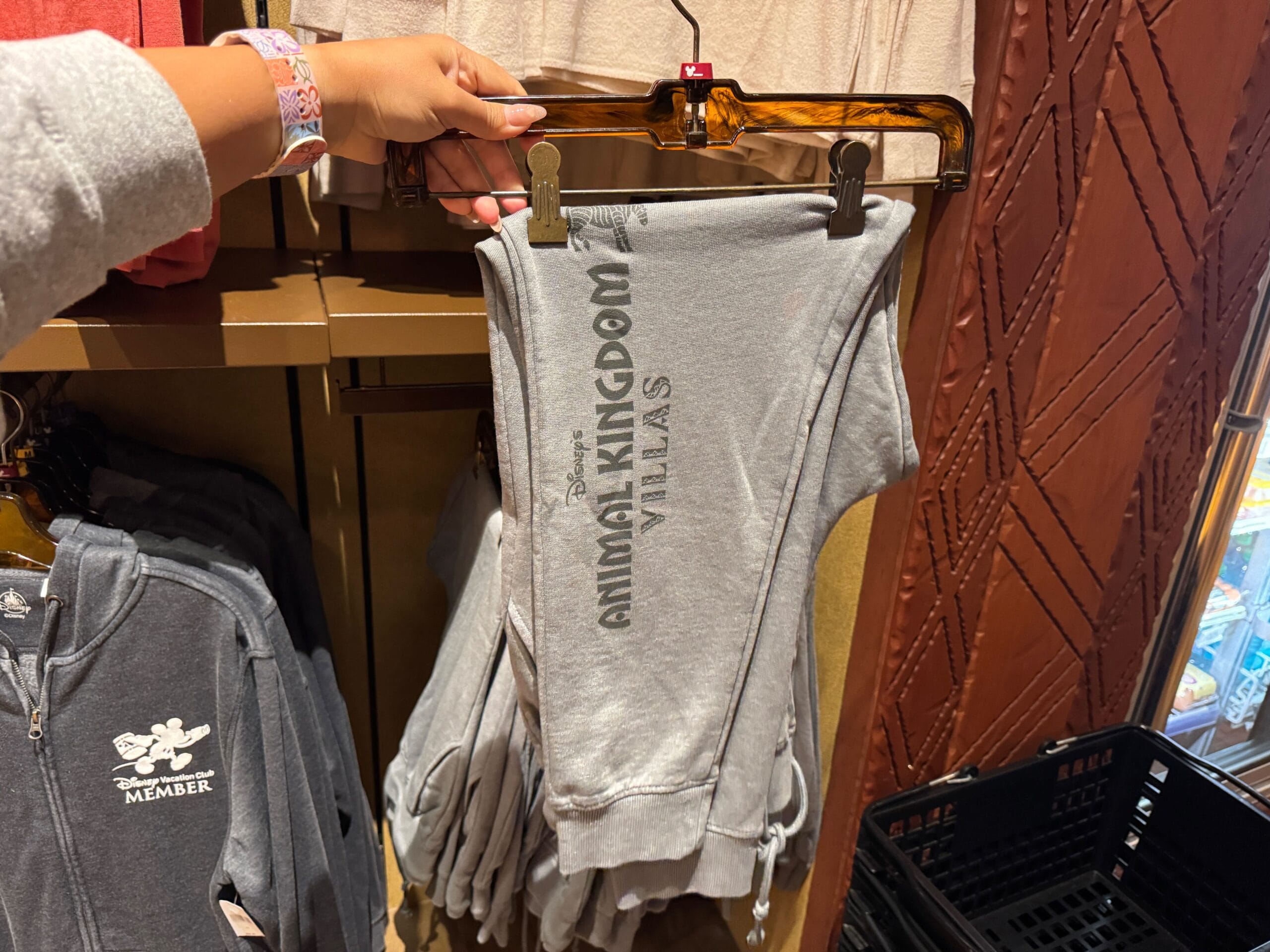 A hand holds up gray sweatpants with "Animal Kingdom Villas" text, displayed on a clothing rack in a store.