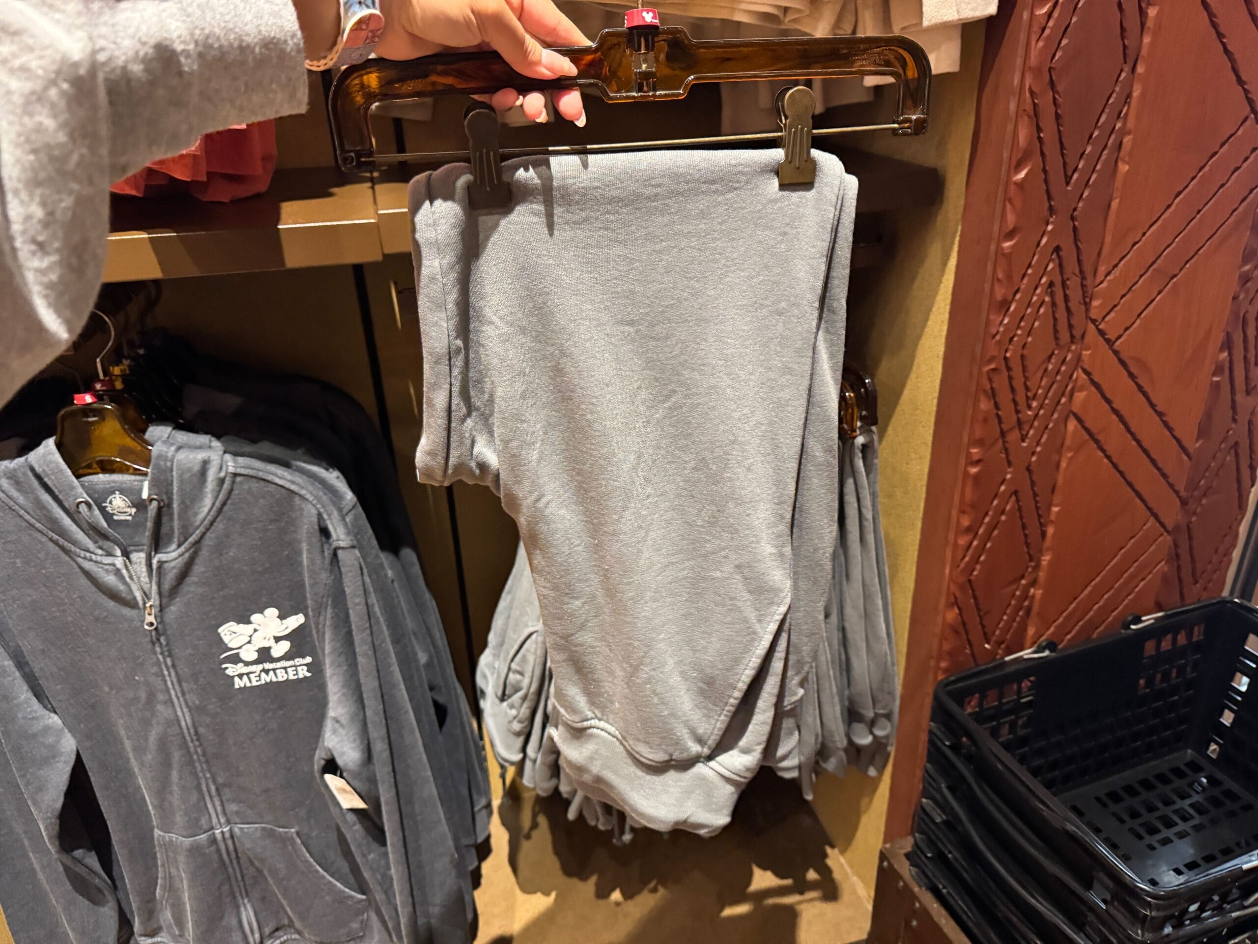 A hand holds gray sweatpants on a hanger; gray hoodies hang nearby on a clothing rack.