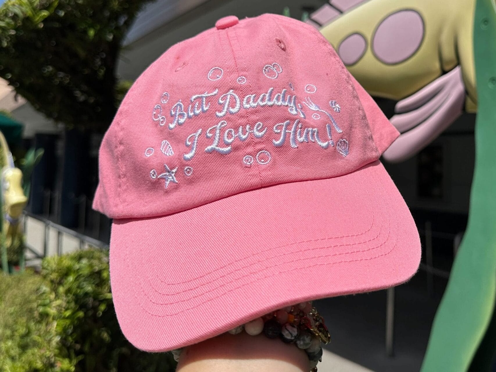 A pink Little Mermaid hat with "But Daddy, I Love Him!" held up by a fish statue and greenery at Disney World.