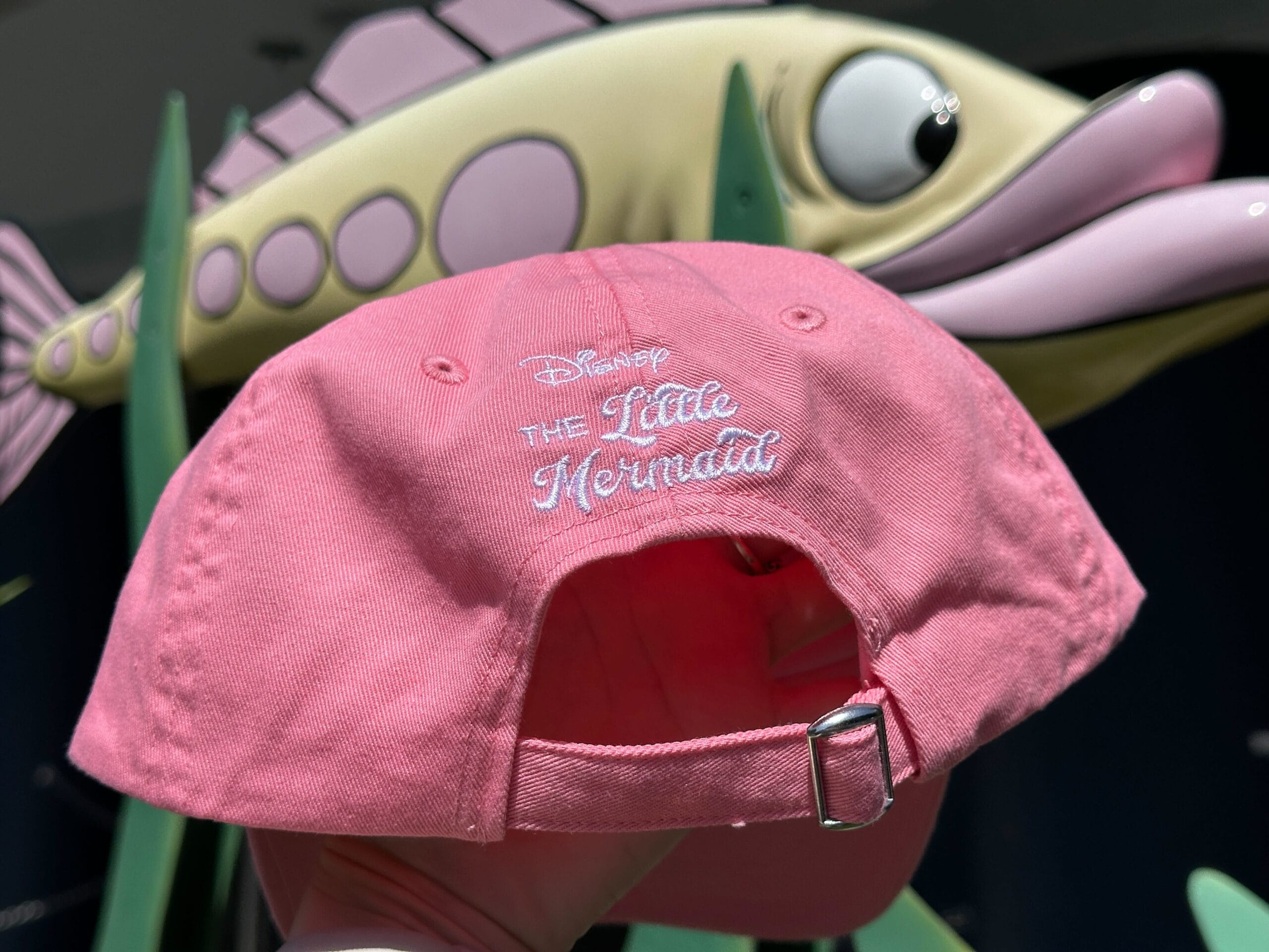 A pink Little Mermaid Hat is held up in front of a fish statue with big eyes and a smiling face.