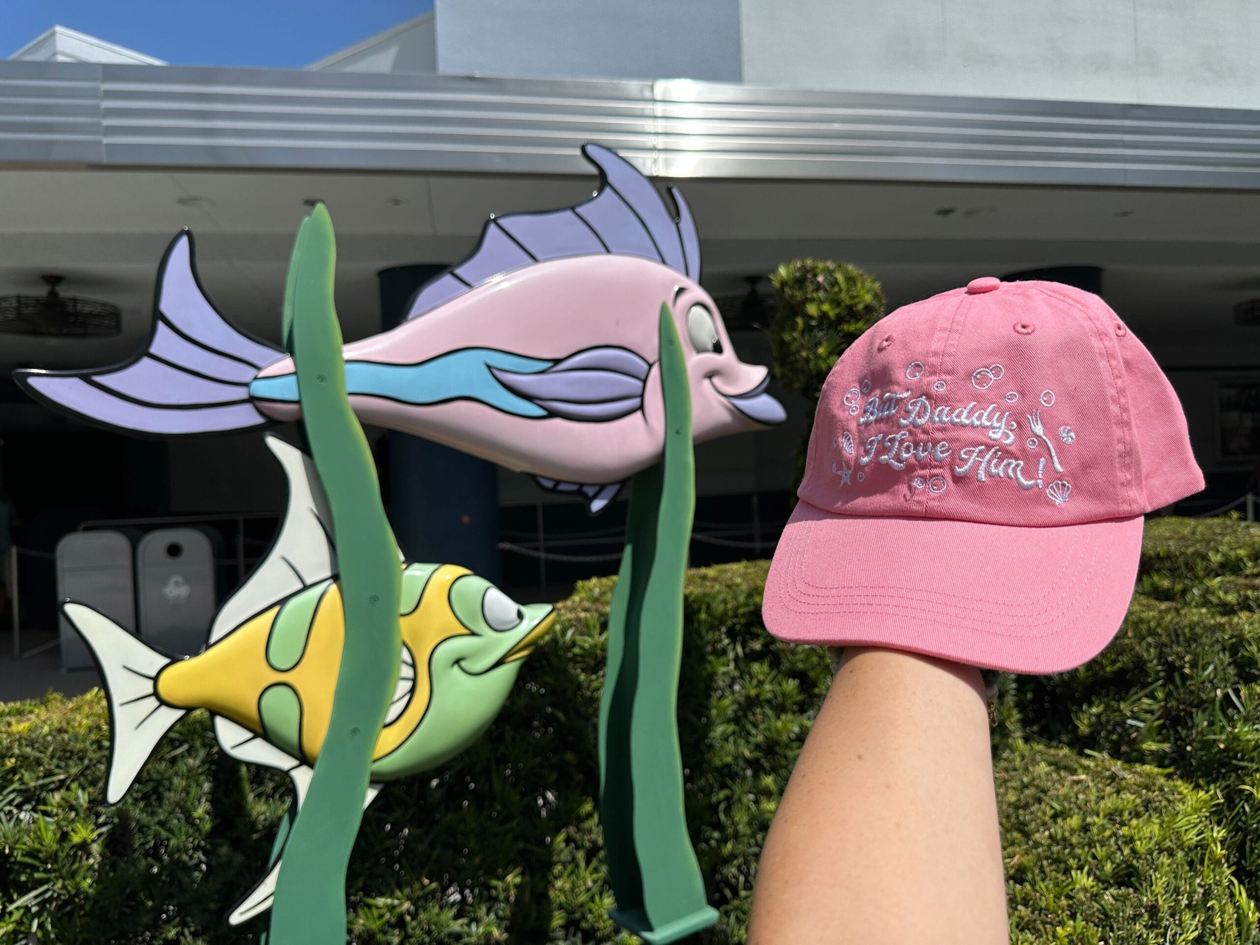 A pink cap reading "Daddy I Love Him!"—a new hat inspired by The Little Mermaid—held before cartoon fish outdoors.