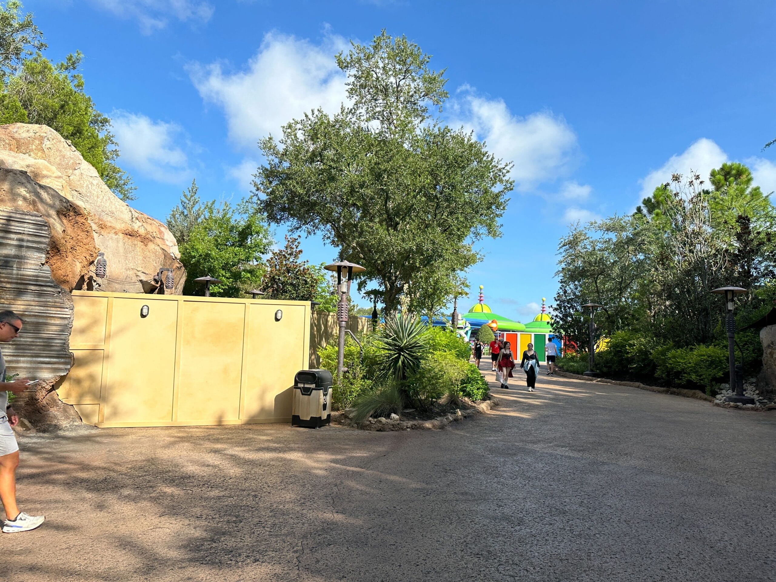 Sunny path with trees, new construction walls on the left, and colorful buildings ahead; a few people walking.