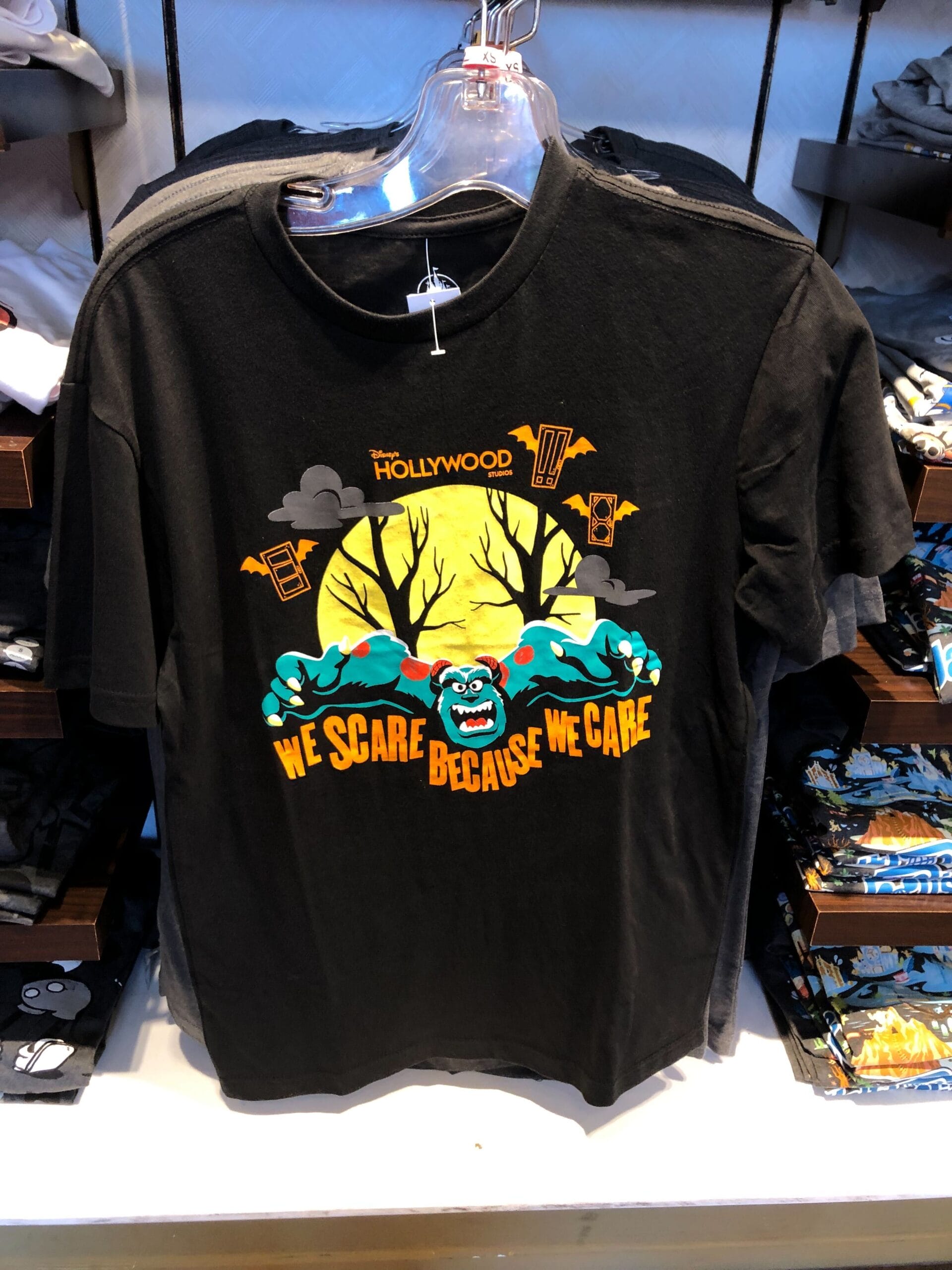 Black T-shirt with a cartoon monster, yellow moon, trees, and text: "We scare because we care" displayed on a rack.