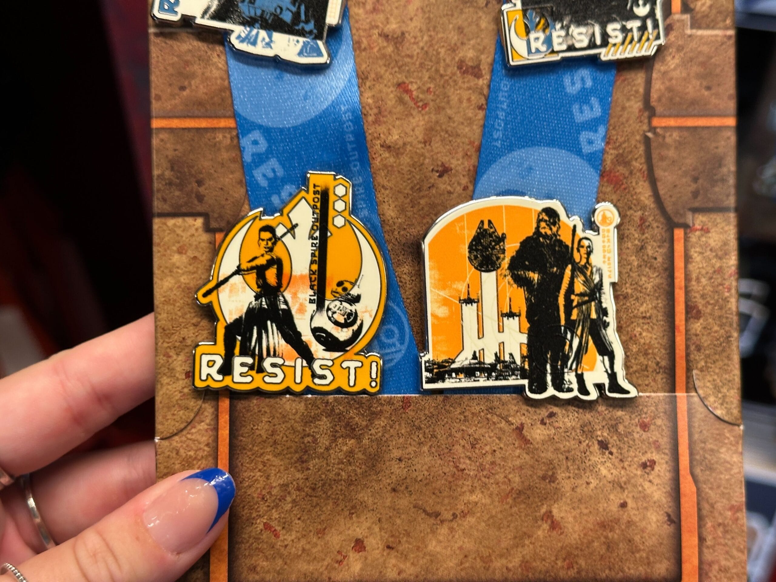 Two "Resist" pins with Star Wars characters on orange backgrounds, shown on a brown board, held by a hand with blue nails.