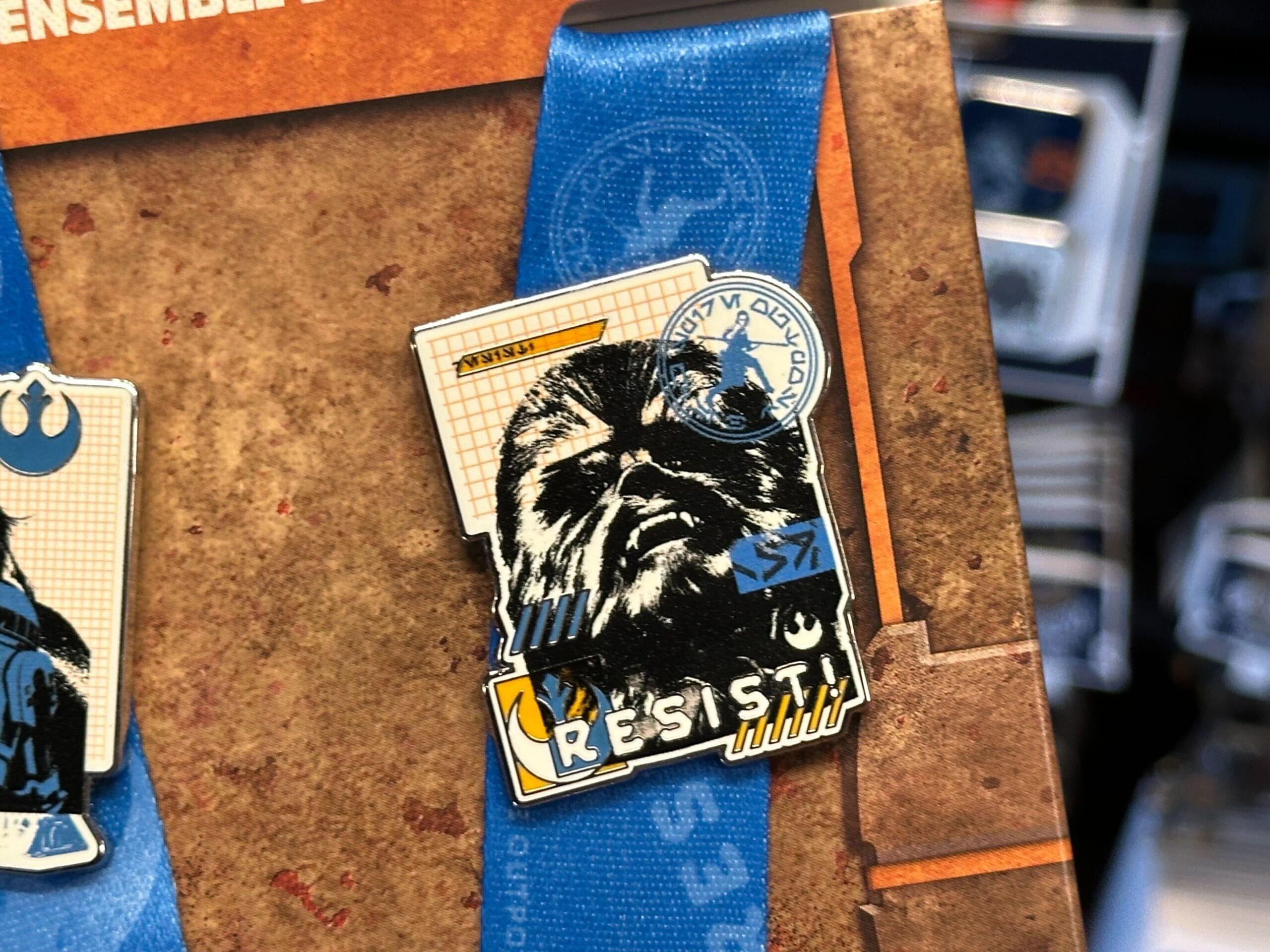 A Chewbacca pin with "RESIST" and a blue ribbon, displayed on a board with other Star Wars pins.