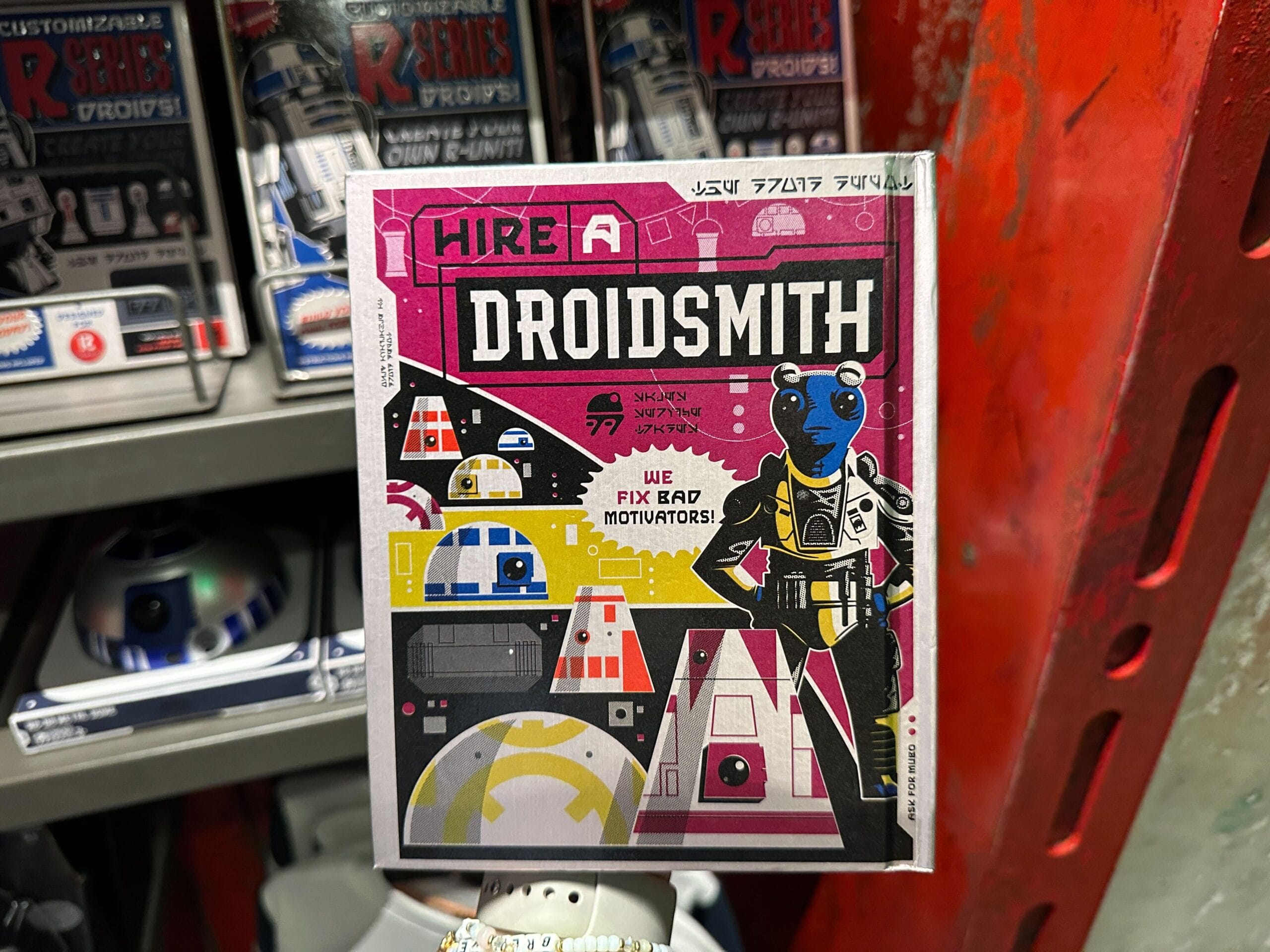 Colorful sign reads “Hire a Droidsmith! We fix bad motivators!” with robots and an alien character illustrated.