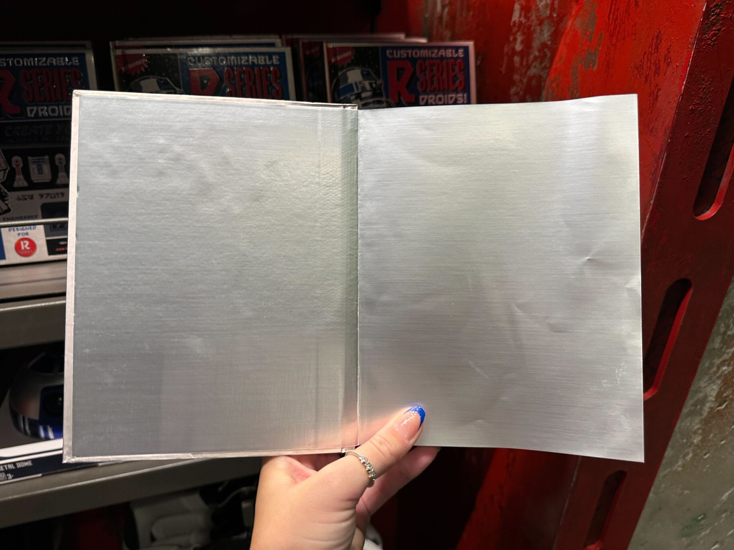 A hand holds open a blank metallic notebook in front of red shelves with toy boxes in the background.