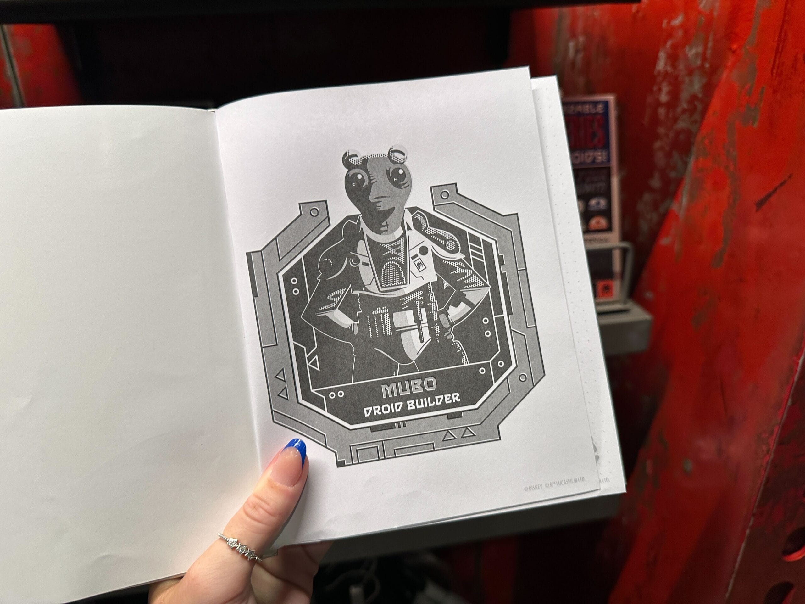 A hand holds open a book showing a drawing labeled "Mubo Droid Builder" against a red background.