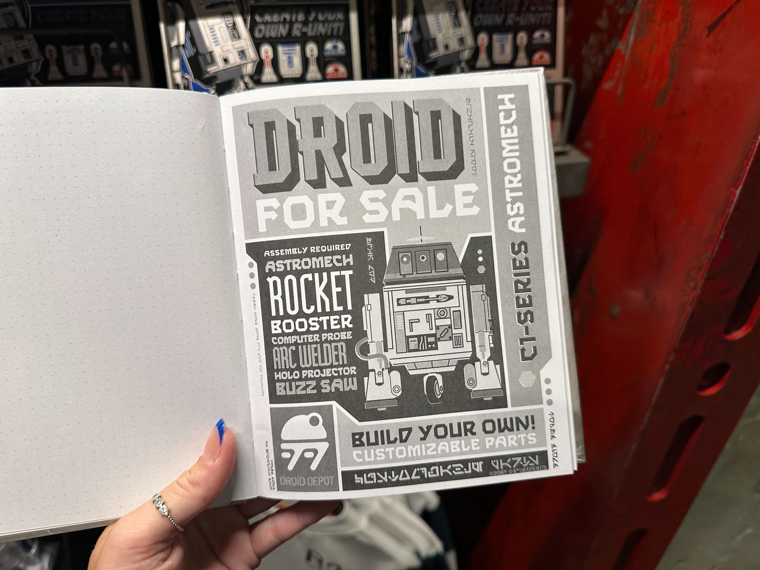 A hand holds an open book showing a “Droid For Sale” advertisement with a cartoon astromech robot and details.