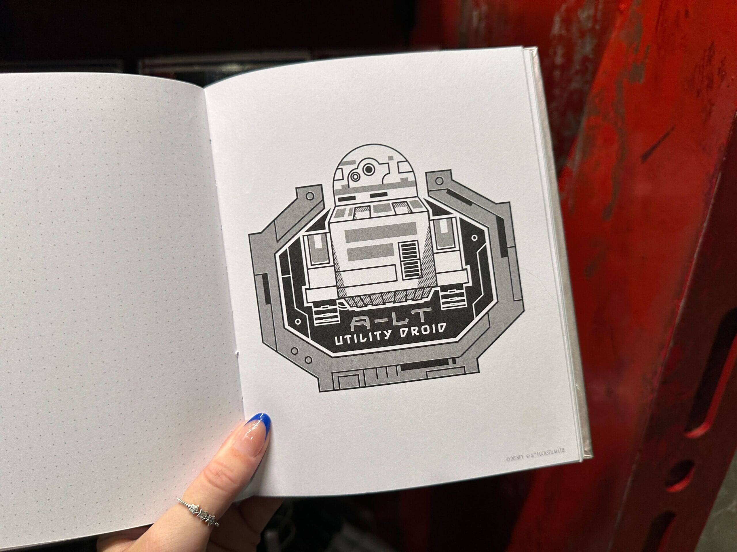 A hand holds an open notebook displaying a black-and-white drawing of a droid labeled "B-LT Utility Droid.