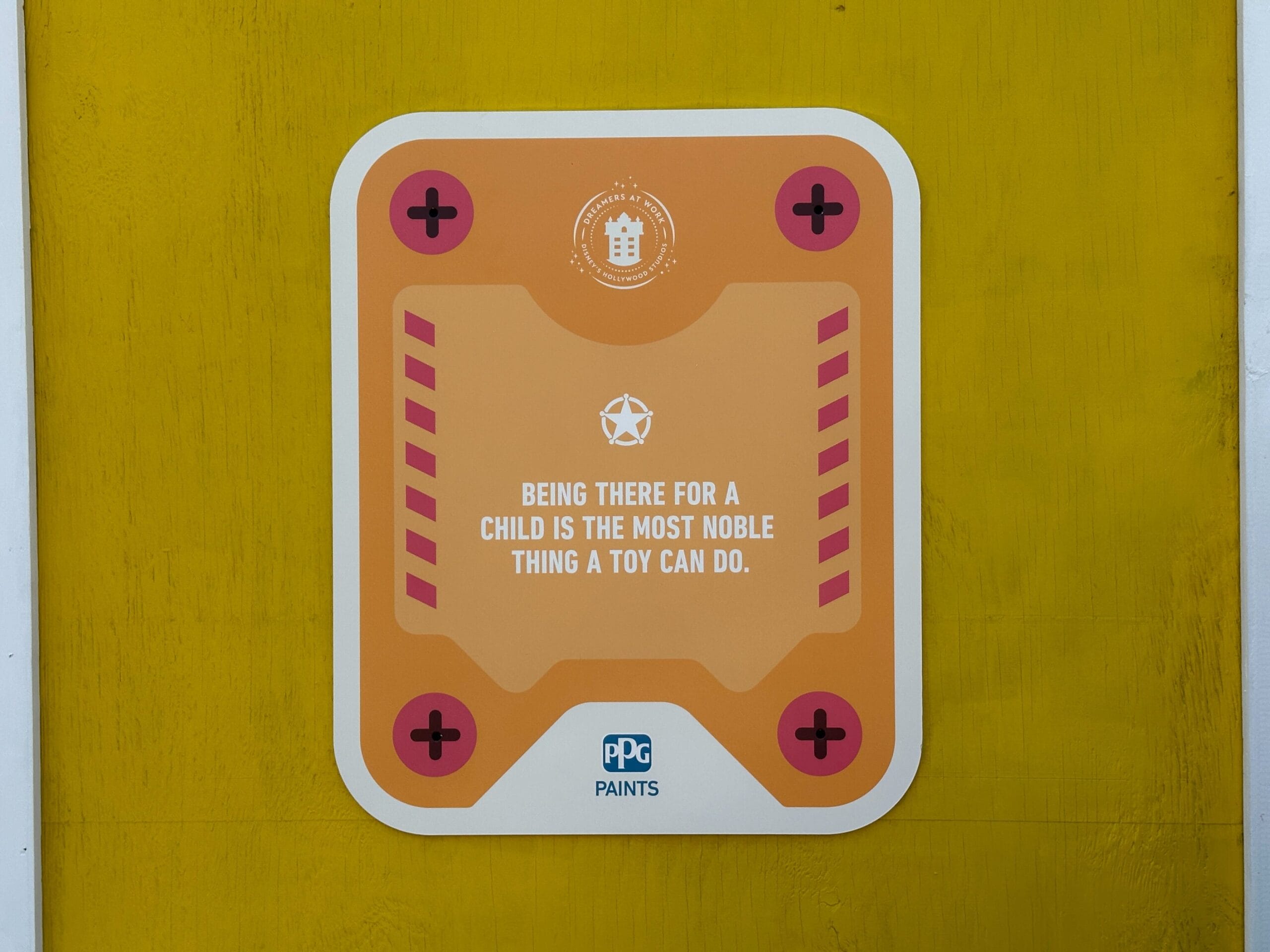 Orange sign on yellow wall reads: "Being there for a child is the most noble thing a toy can do." Toy Story wall art style.