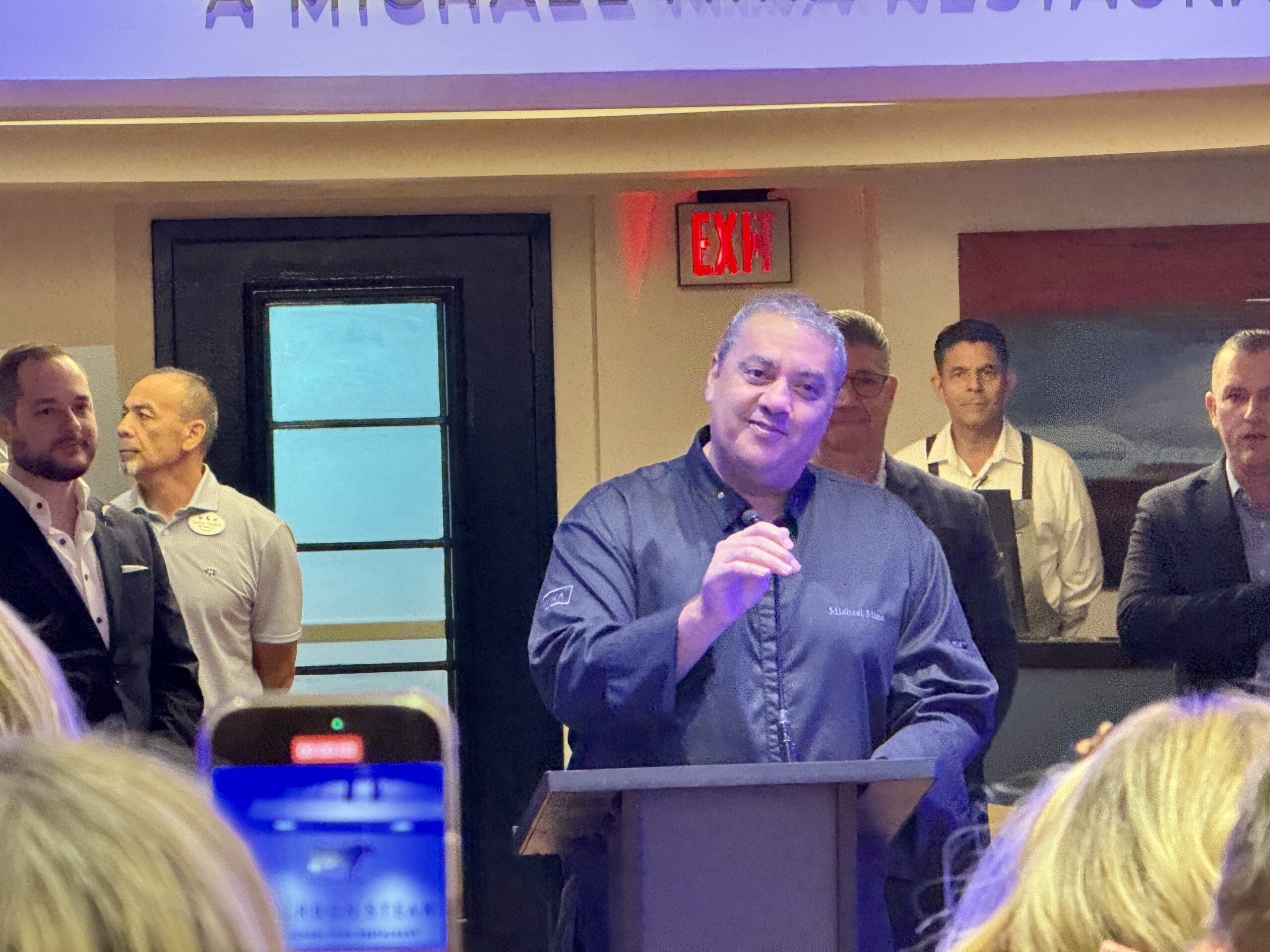 A chef speaks at a podium in a restaurant, like Michael Mina at Bourbon Steak, as staff and guests snap photos.