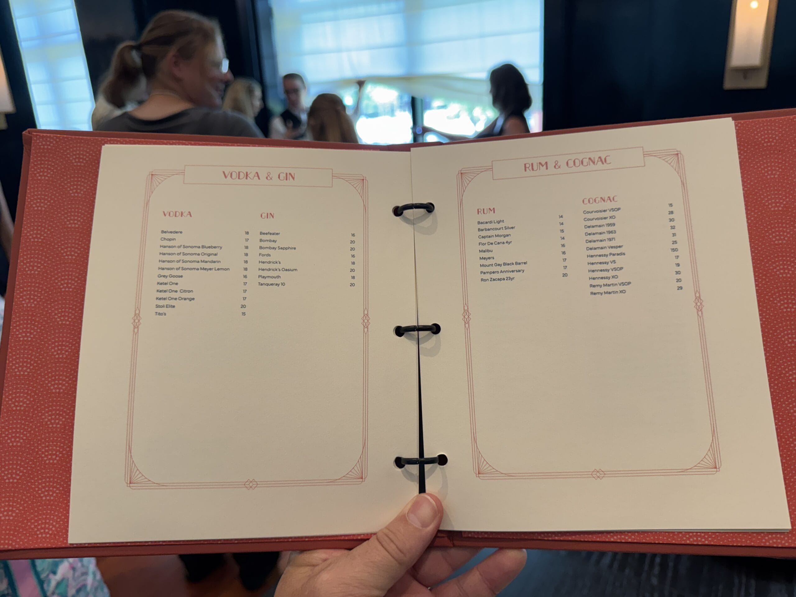 A hand holds open a drink menu at Bourbon Steak, Michael Mina’s spot in Walt Disney World Dolphin Hotel; people mingle behind.