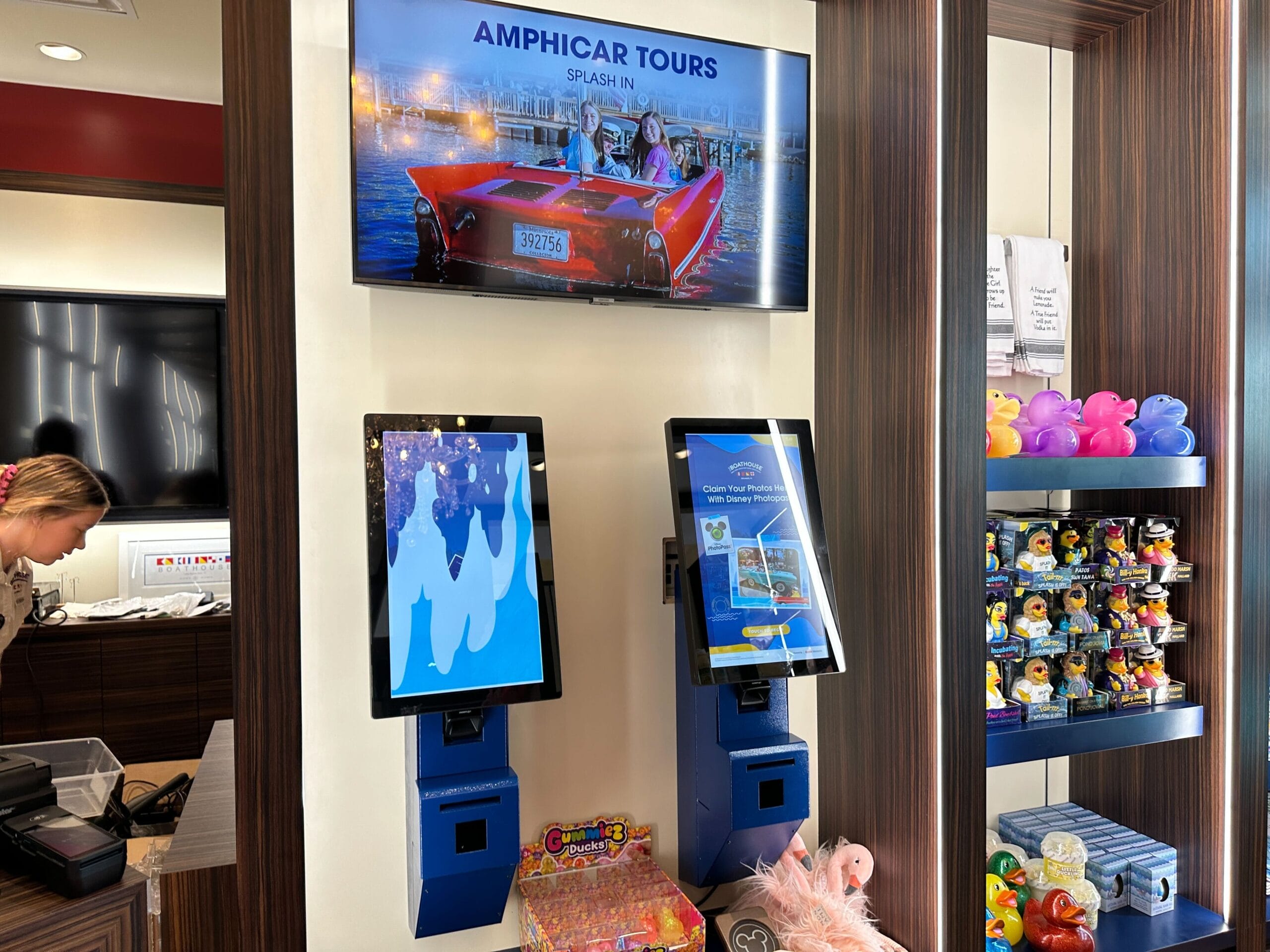 Two touchscreen kiosks beneath an Amphicar Tours sign, with rubber ducks and toys displayed on shelves nearby.
