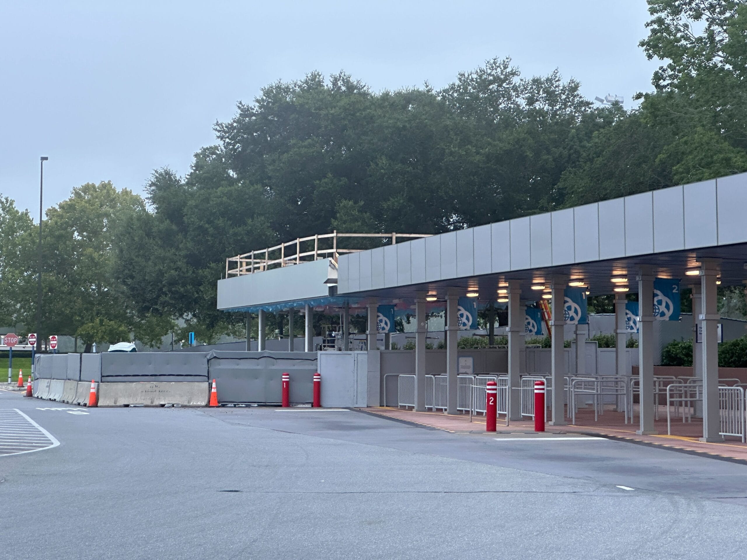 A mostly empty queue area with construction barriers and cones near a covered walkway, surrounded by trees.