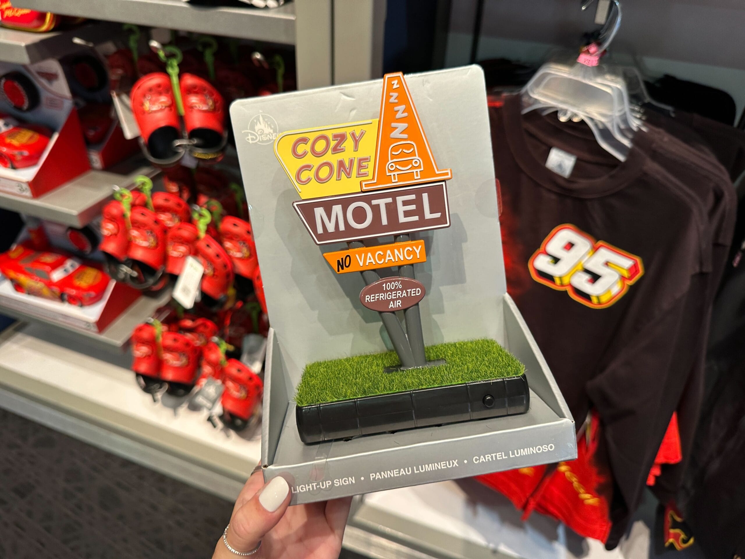 Hand holding a “Cozy Cone Motel” light-up sign from Disney’s Cars, displayed near Cars-themed merchandise.