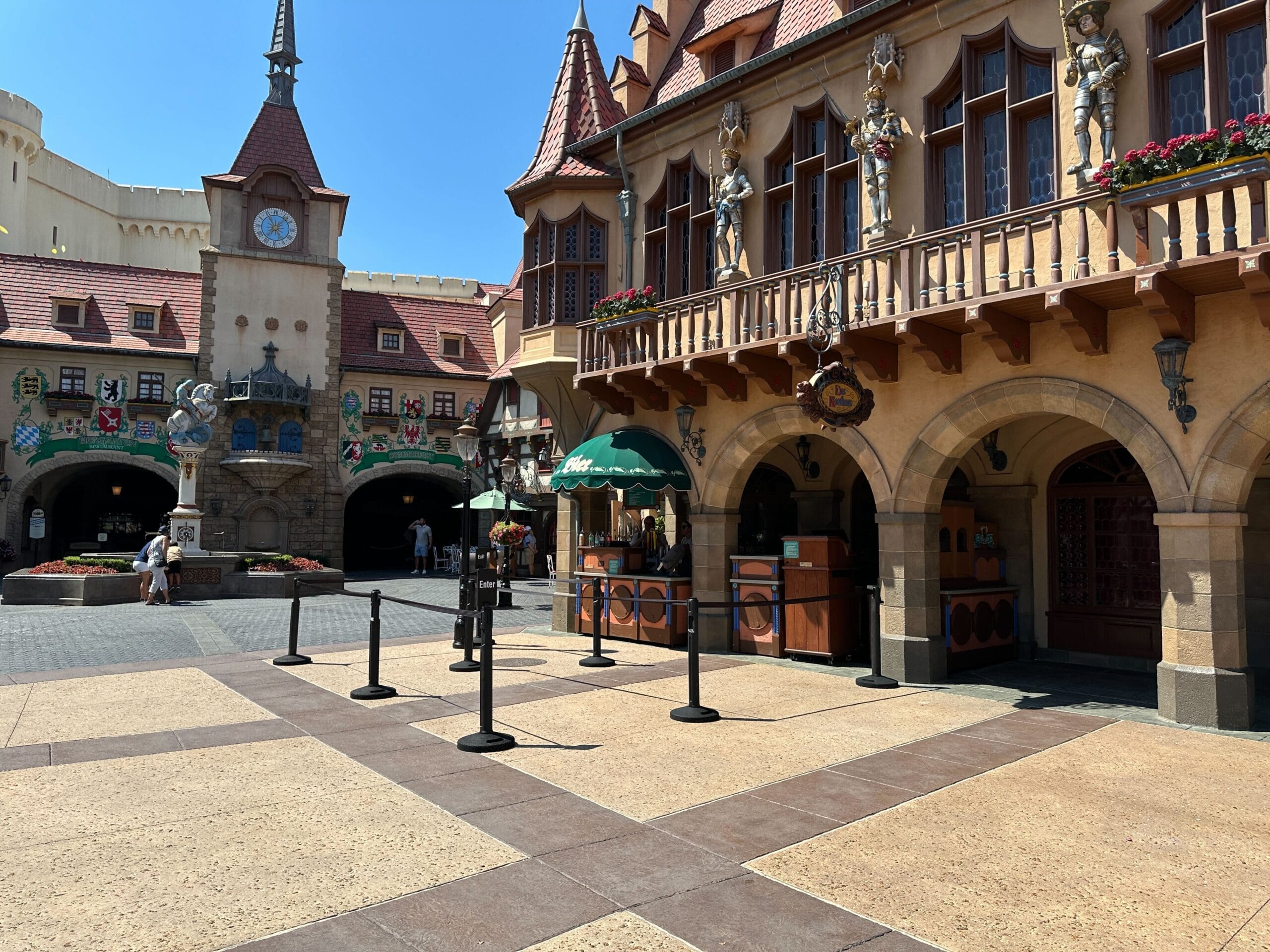 German-style plaza with decorative buildings, clock tower, and a shaded food stand on a sunny day.