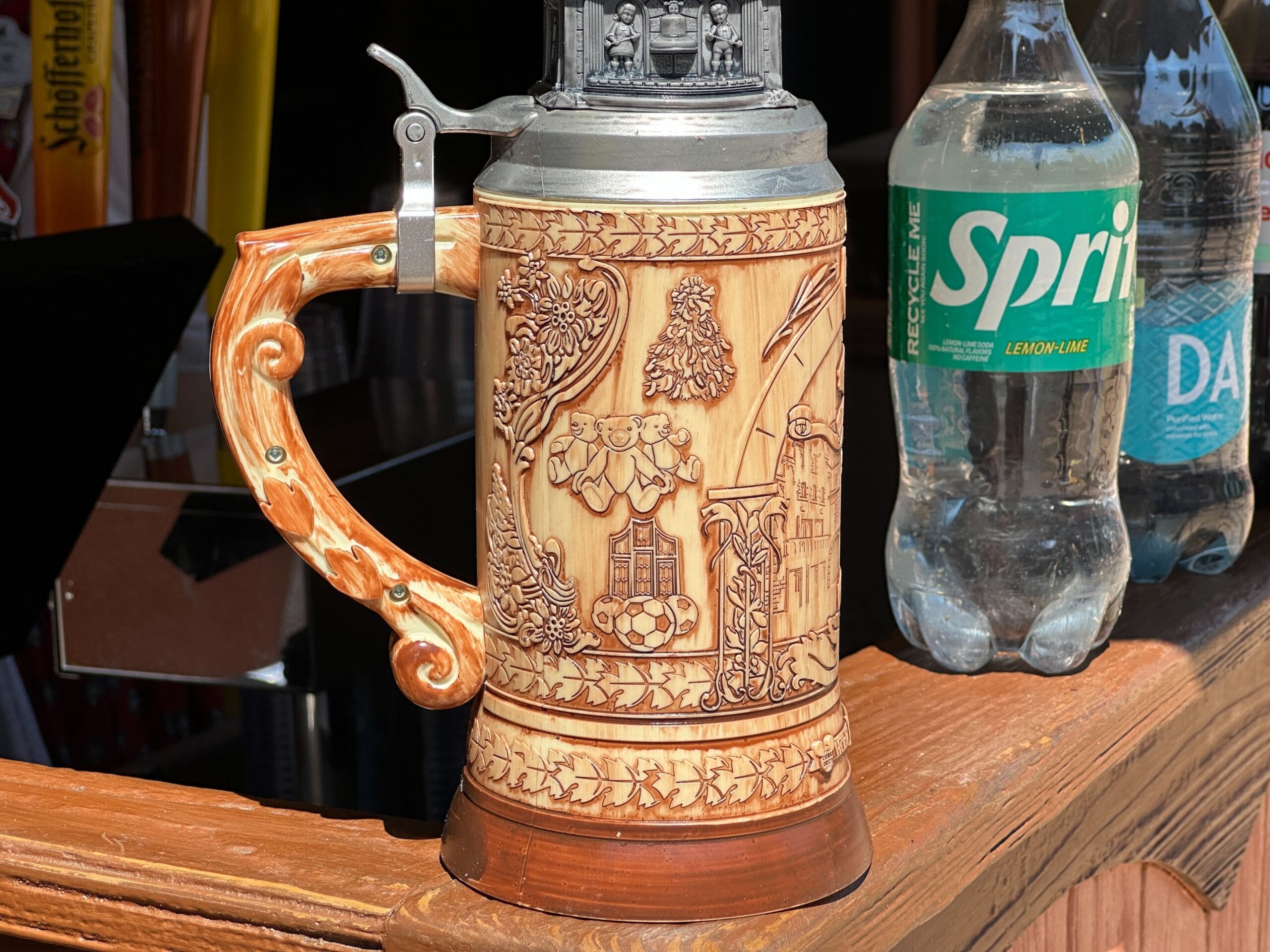 A decorative beer stein with an ornate handle beside bottles of Sprite and water on a wooden counter.