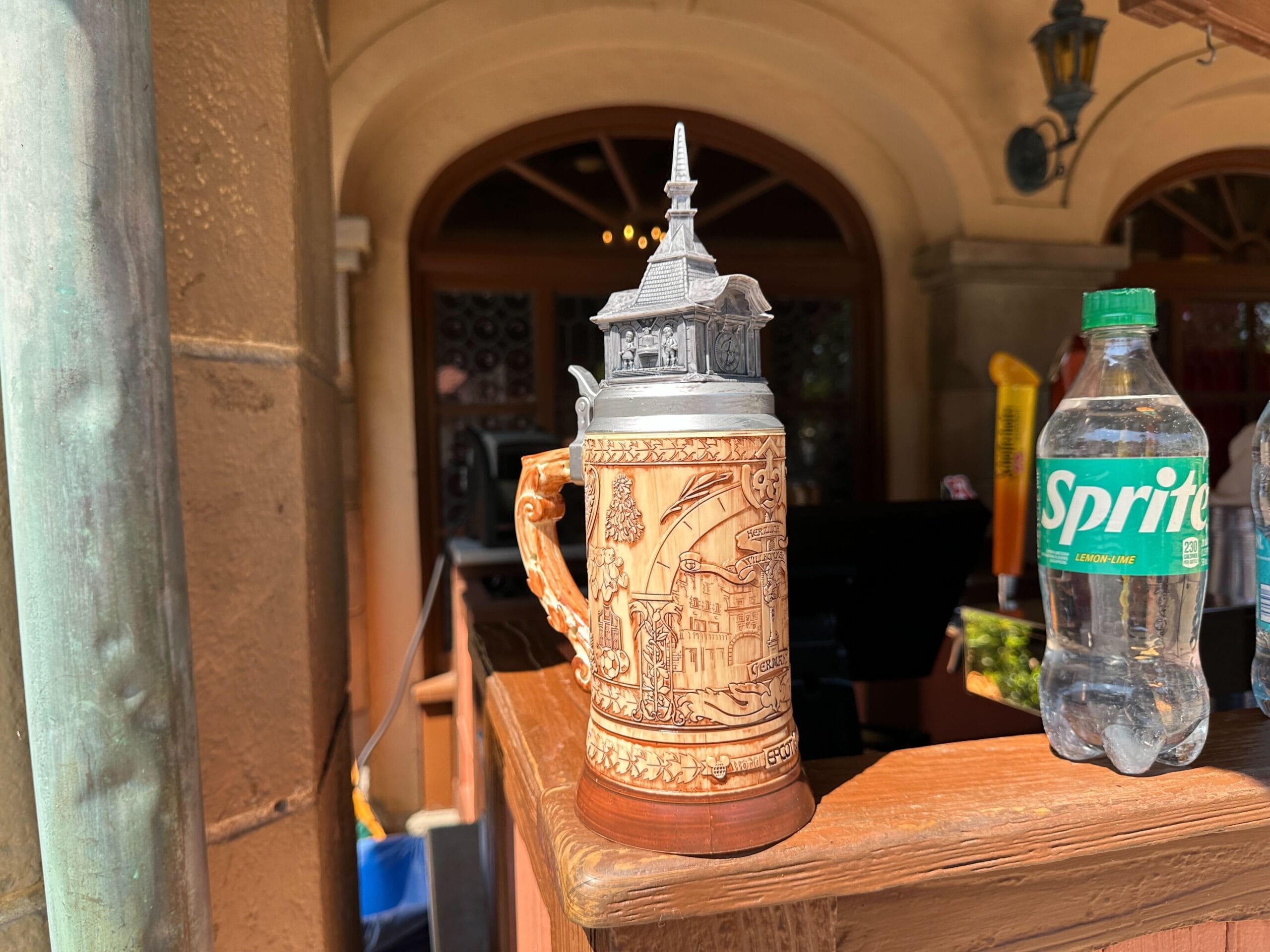 A decorative beer stein sits on a wooden counter next to a bottle of Sprite in an outdoor setting.