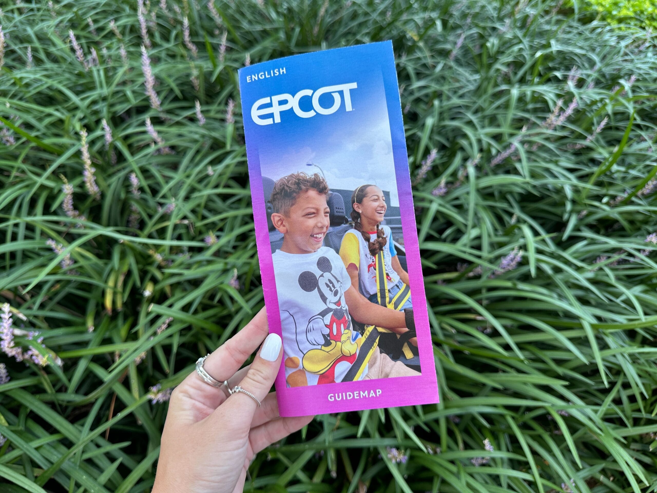 A hand holds a reimagined EPCOT map with smiling kids in Mickey shirts and lush green plants in the background.