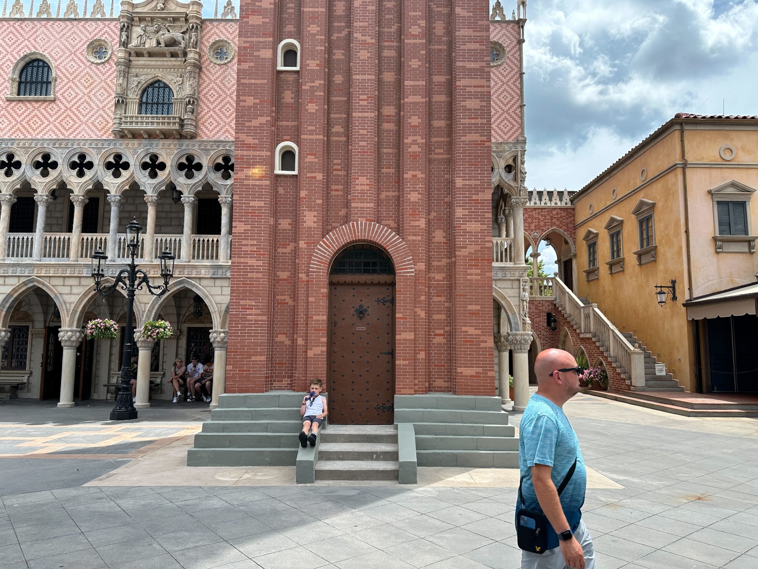 A man walks in front of a red brick building, while a child sits on steps in the background, under a cloudy sky.