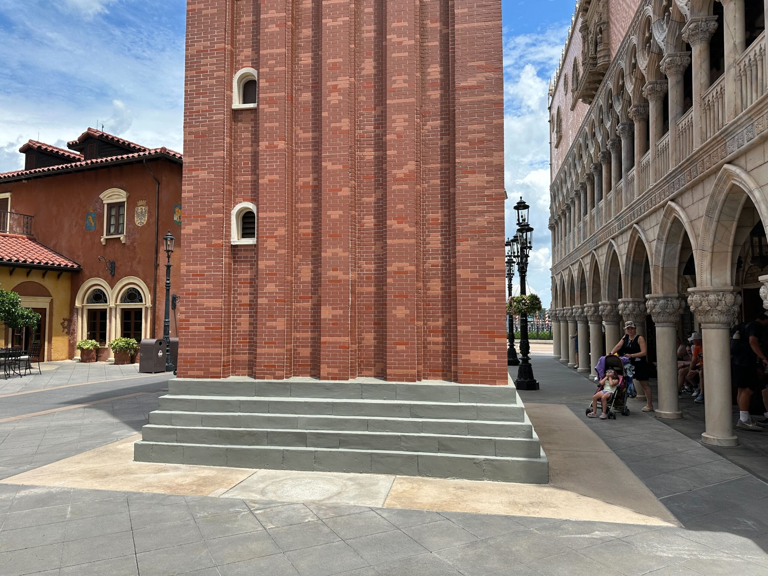 A tall red-brick tower with small windows and gray steps stands in a plaza with people and arched walkways nearby.