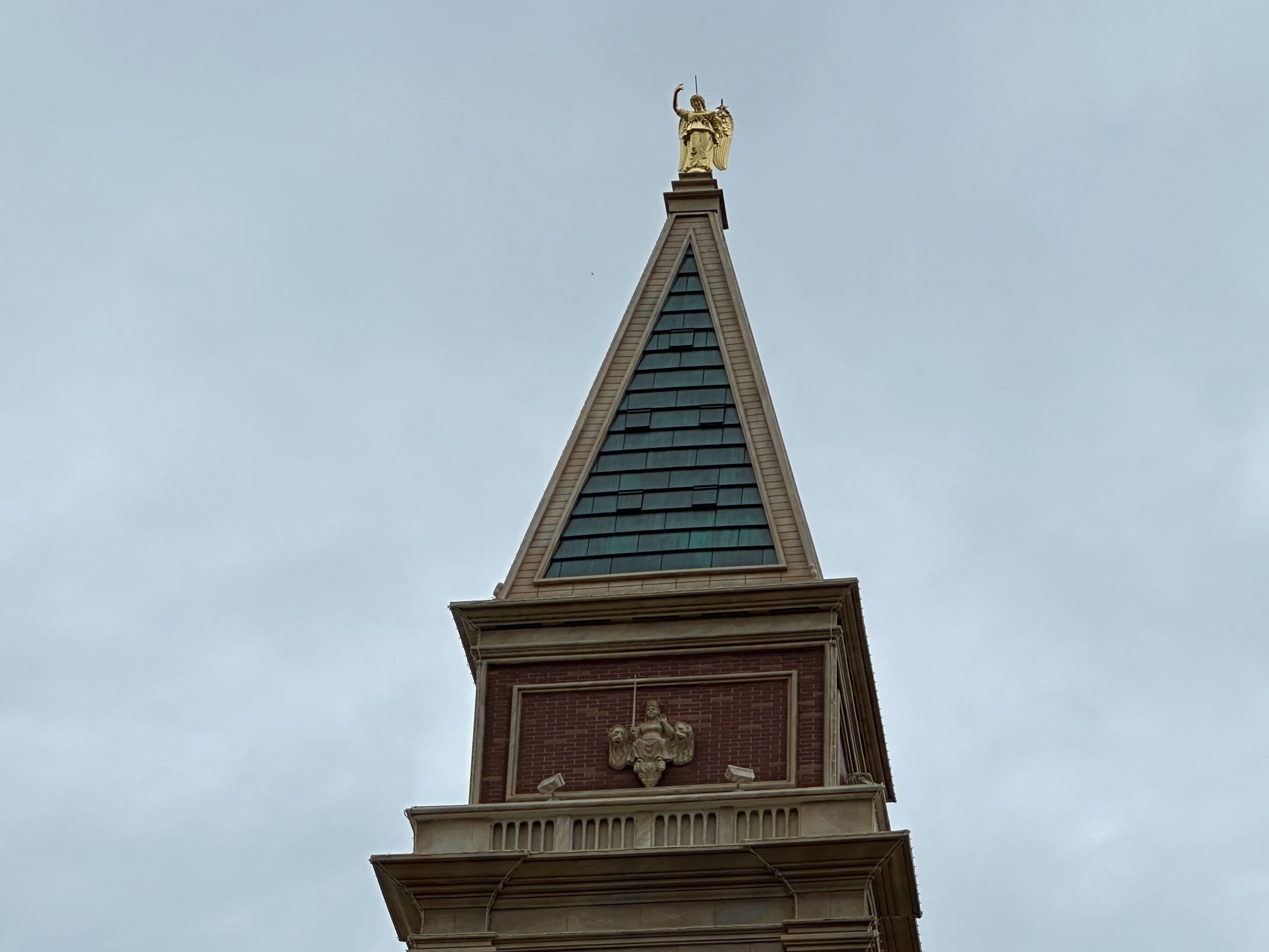 A pointed tower with a gold statue on top against a cloudy sky.