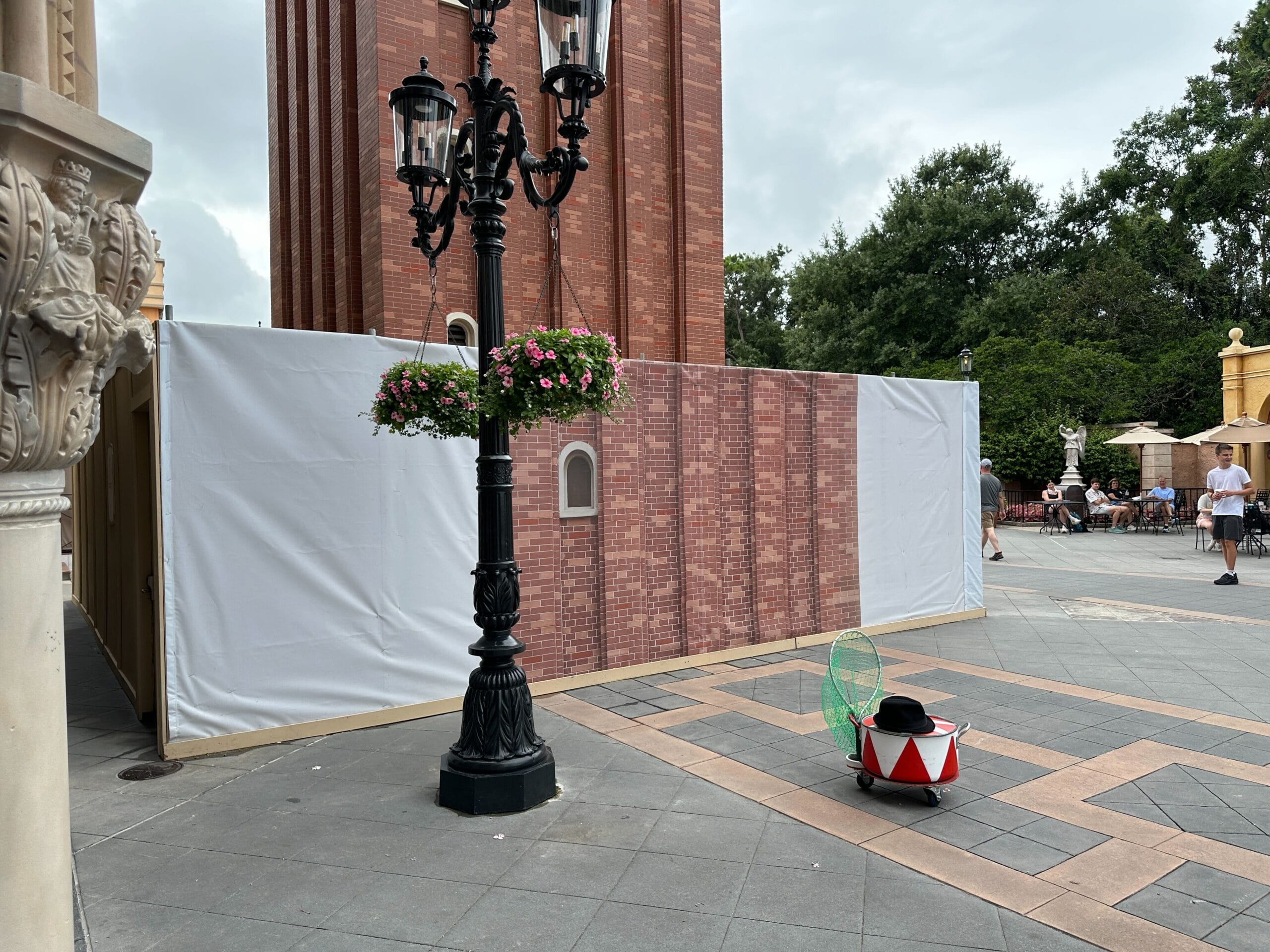 A construction wall with a brick pattern stands near a lamppost and a drum-shaped robot in an outdoor plaza.