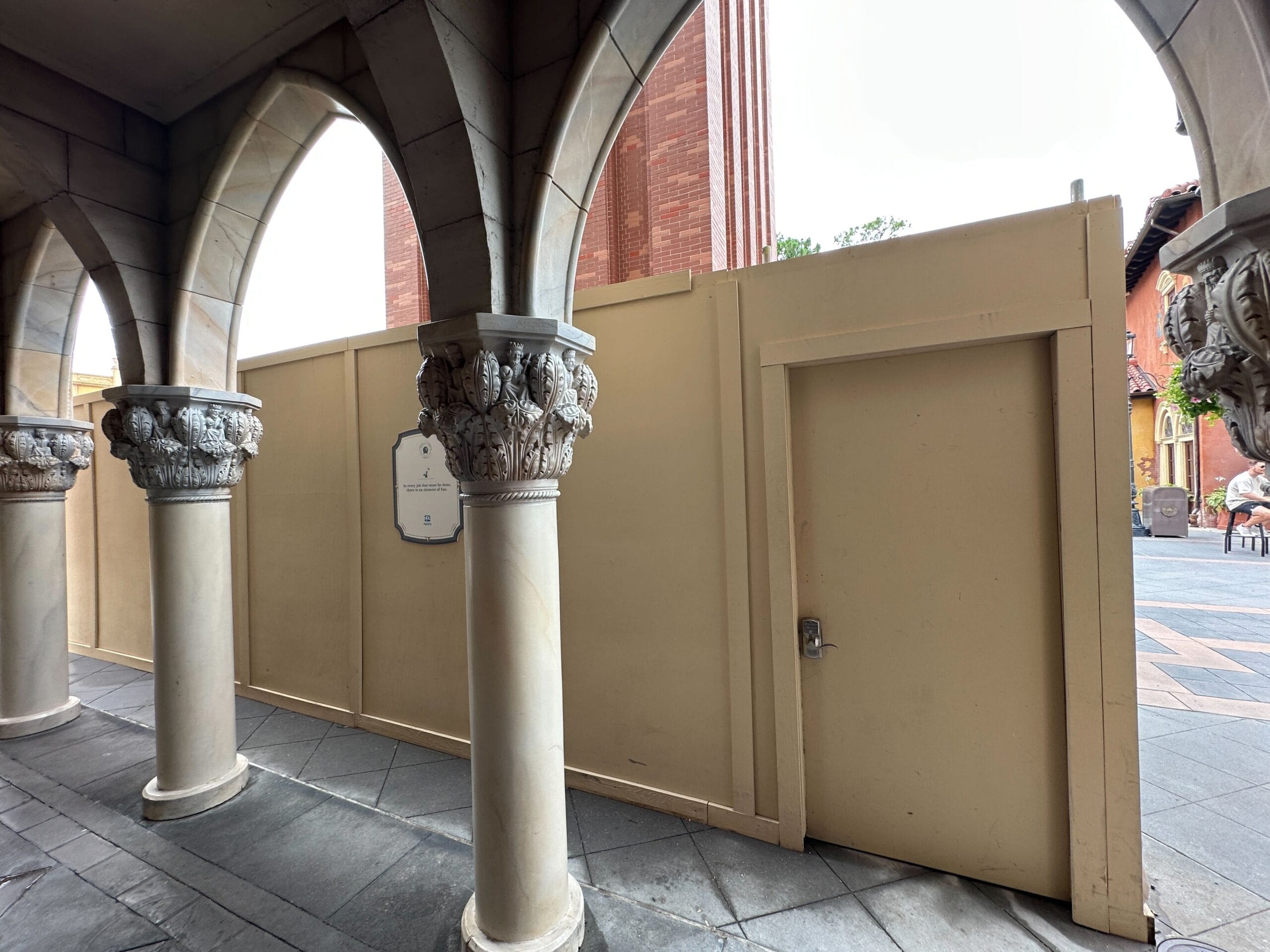 Tan construction walls and door block off an area beside decorative stone columns and arches in an outdoor walkway.