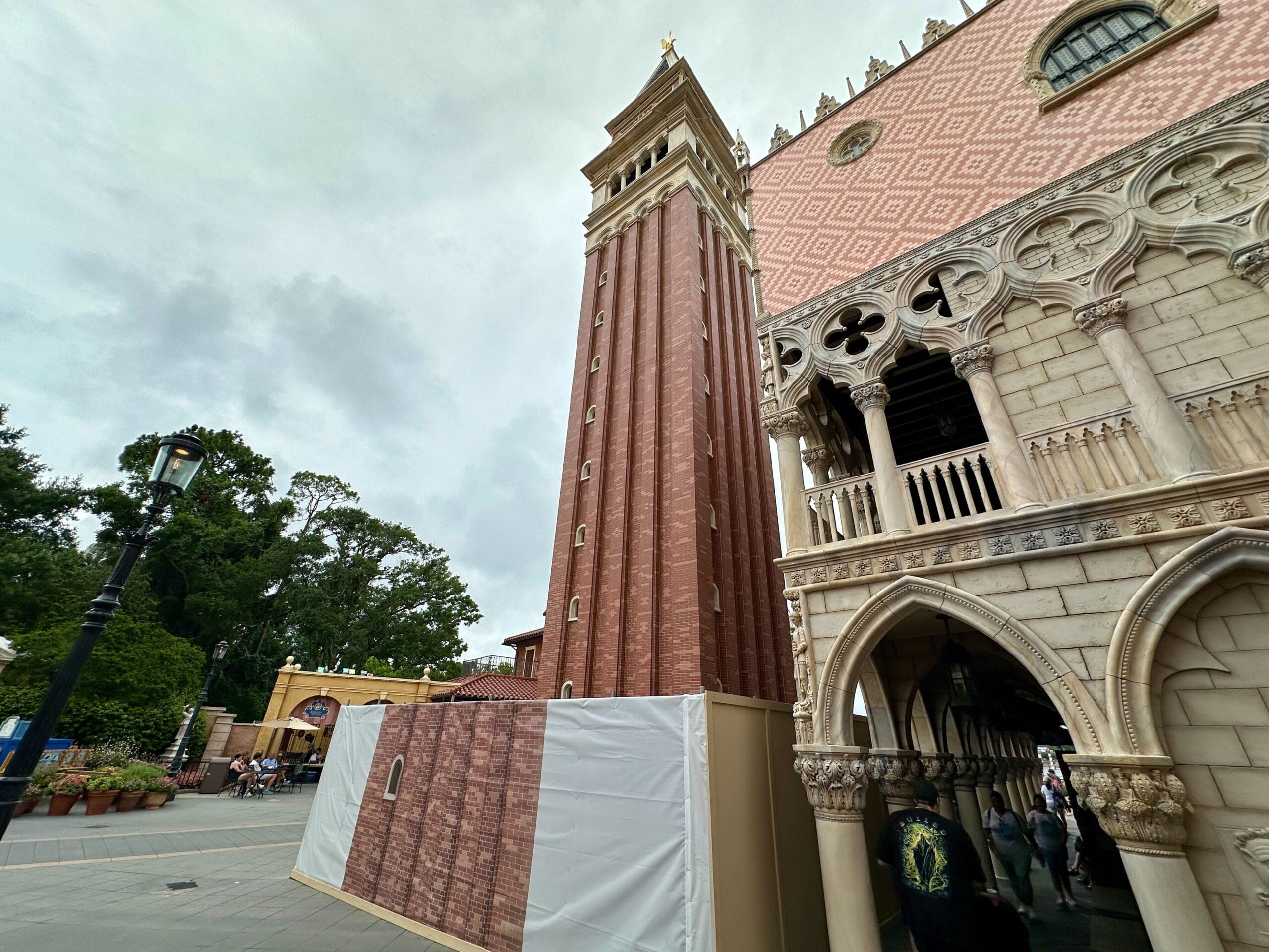 Tall, red-brick bell tower with construction barriers, next to ornate building under a cloudy sky.