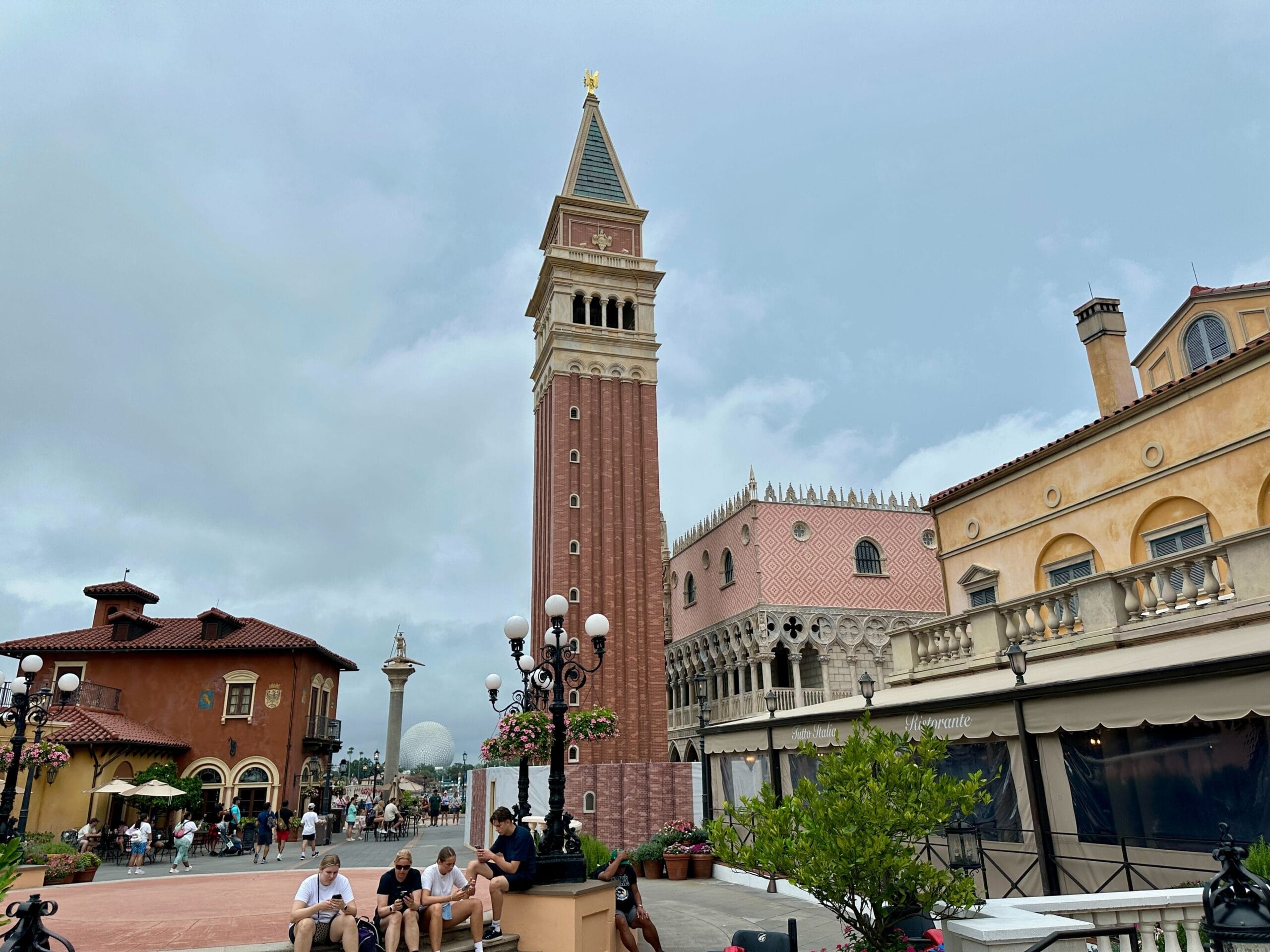 A tall brick bell tower rises among Italian-style buildings with people sitting and walking nearby under a cloudy sky.