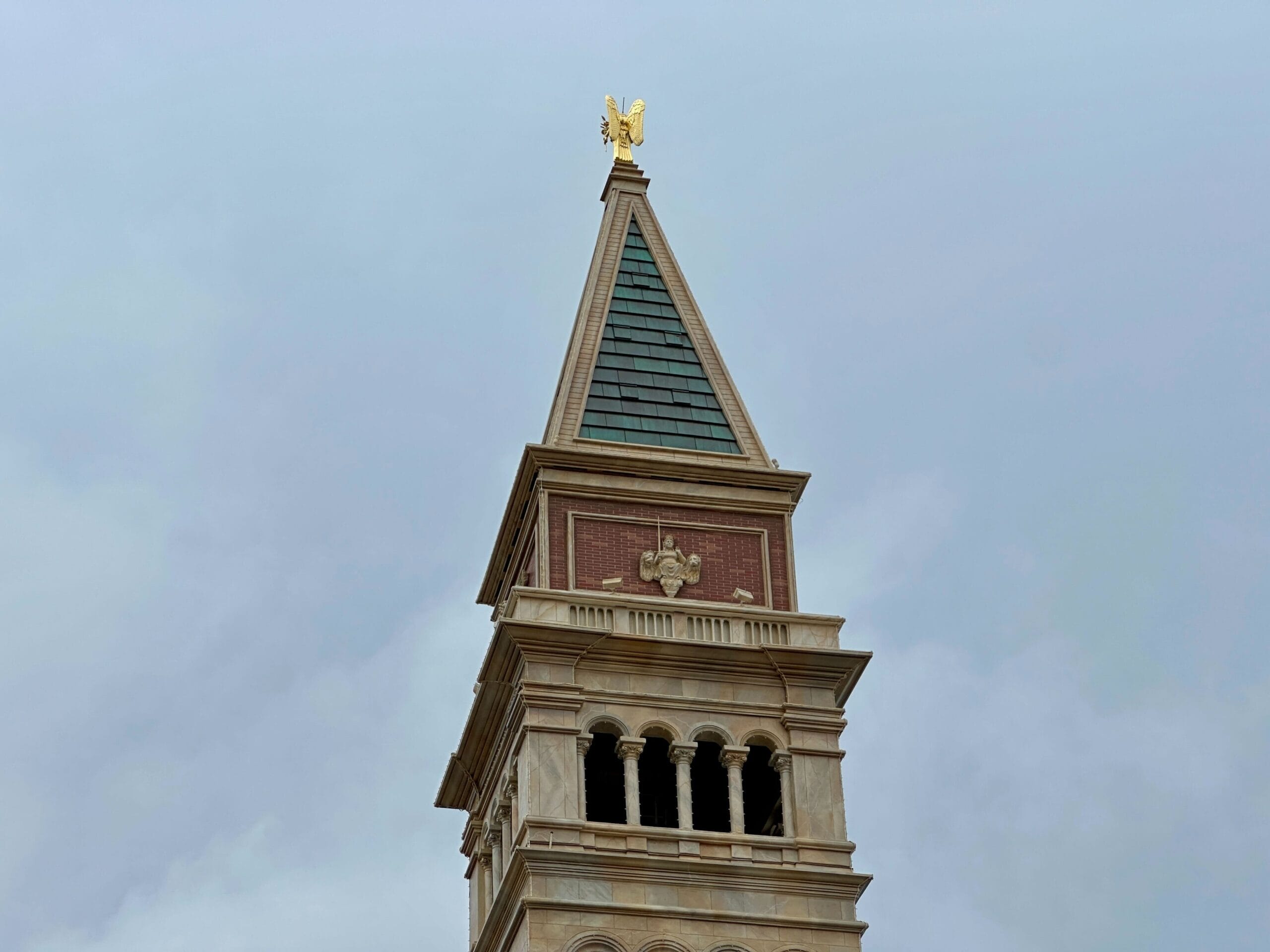 A tall stone clock tower with arched windows and a green-tiled spire topped by a golden statue against a cloudy sky.