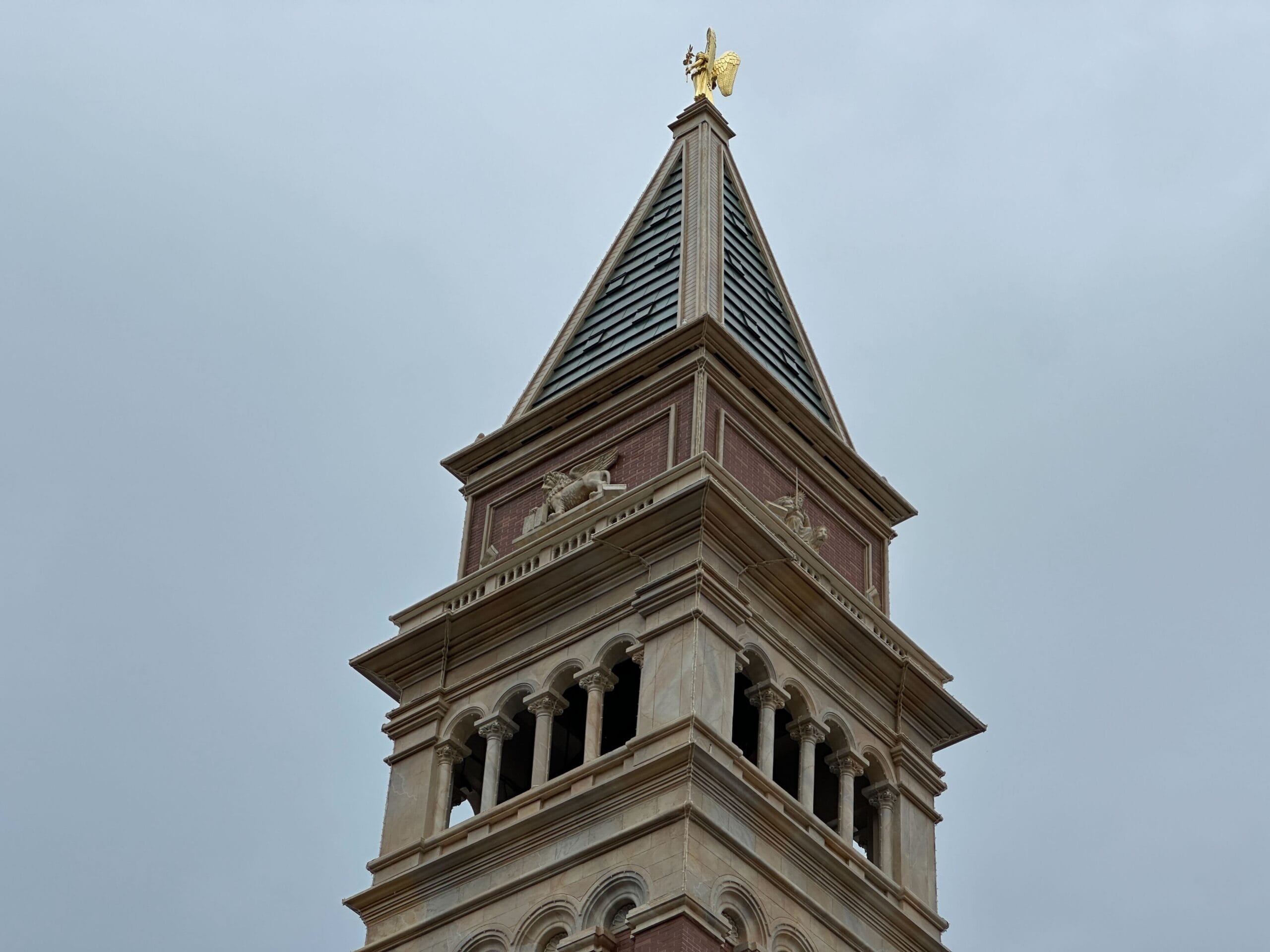 Tall brick clock tower with arched windows and a golden angel statue on top, set against a cloudy sky.