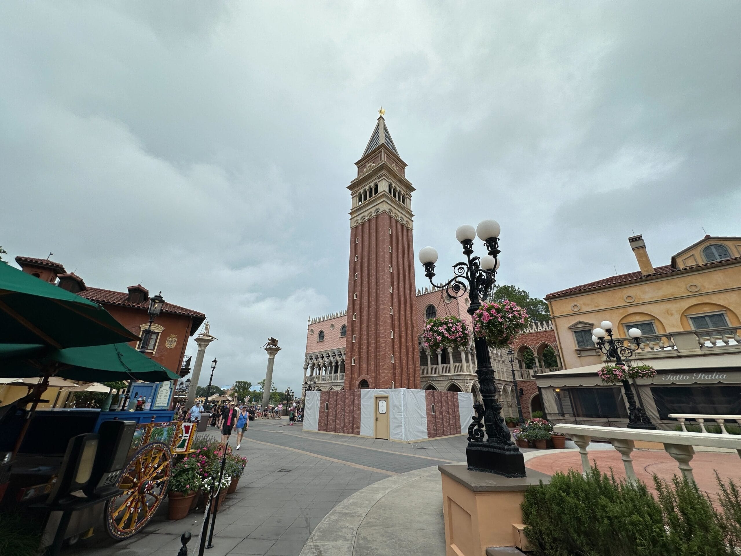 A tall brick bell tower stands in a plaza surrounded by shops and people under a cloudy sky.