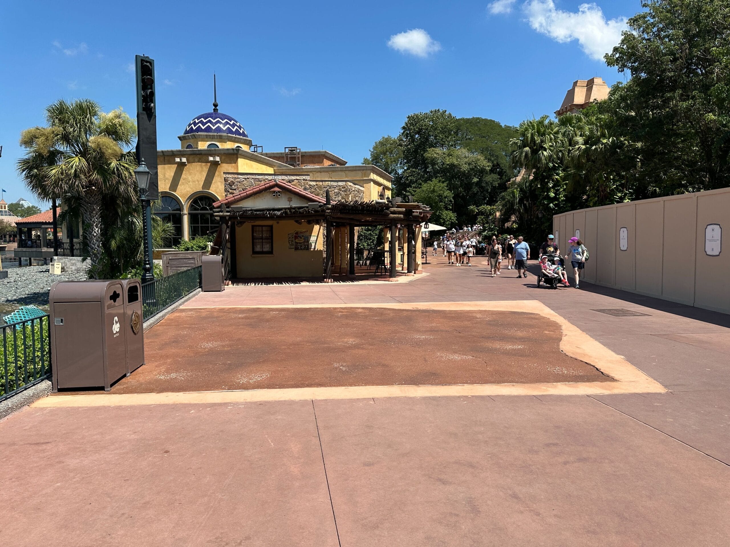 A wide theme park walkway with construction walls and people walking under a sunny, blue sky.