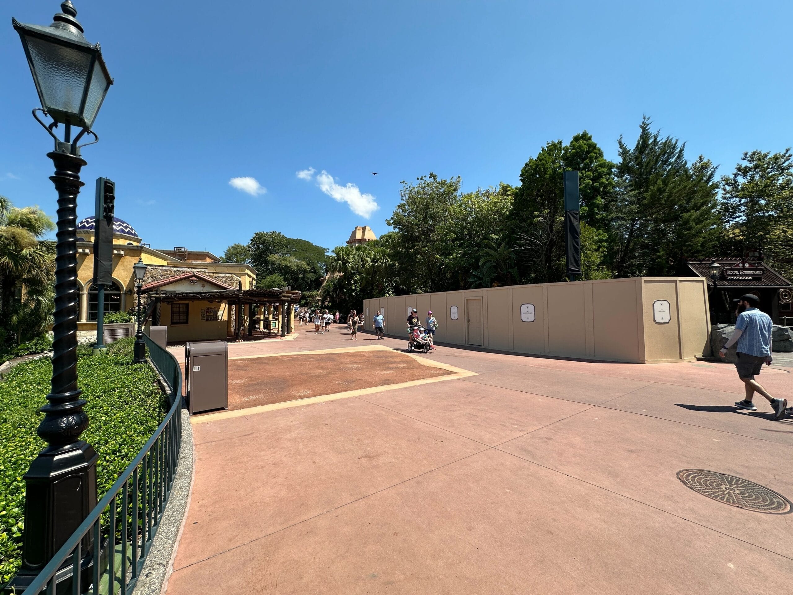 A theme park walkway with construction walls on the right, people walking, and trees under a clear blue sky.