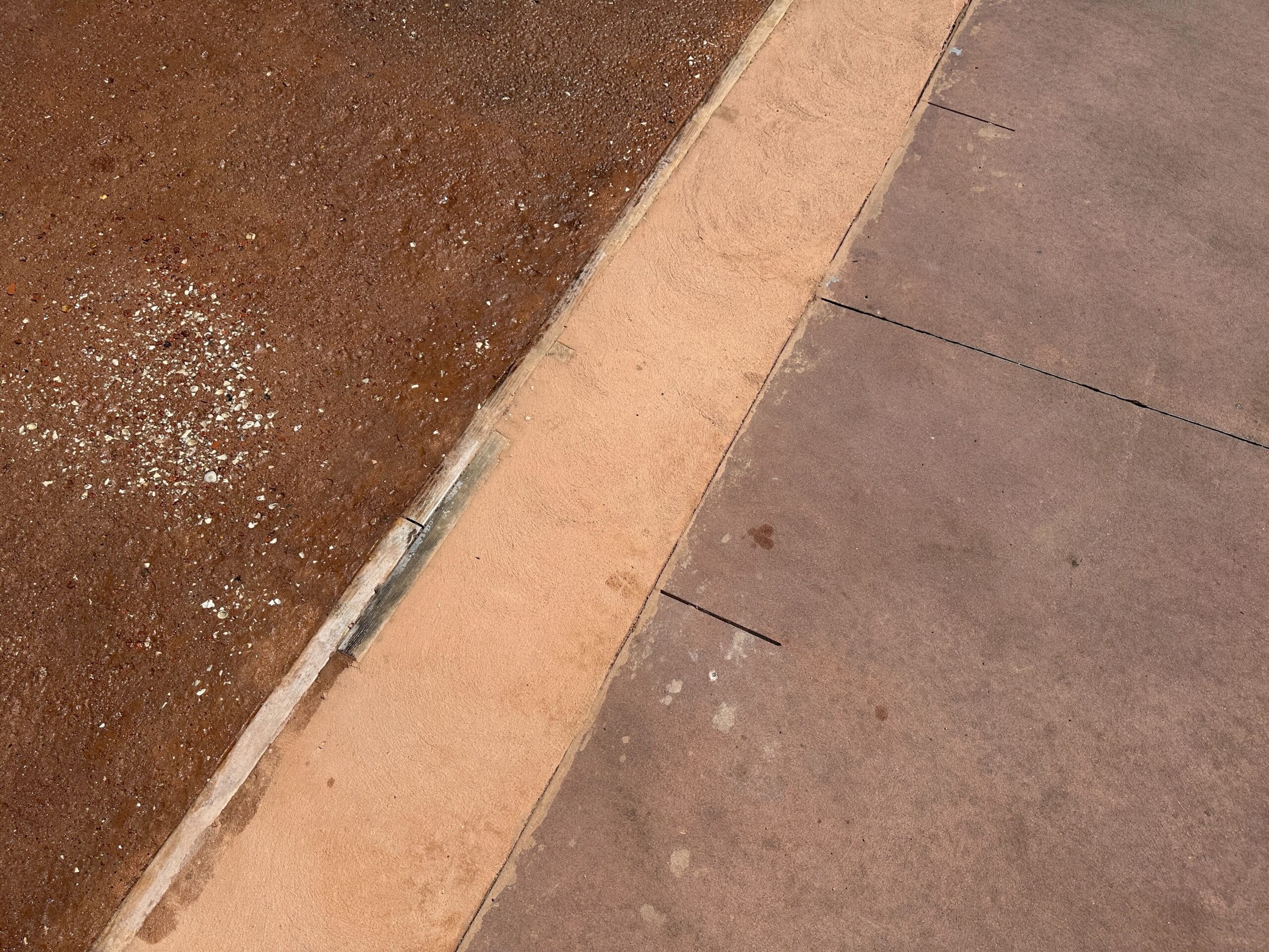A sidewalk meets a gravelly dirt area, separated by a thin, sandy strip and a concrete curb.